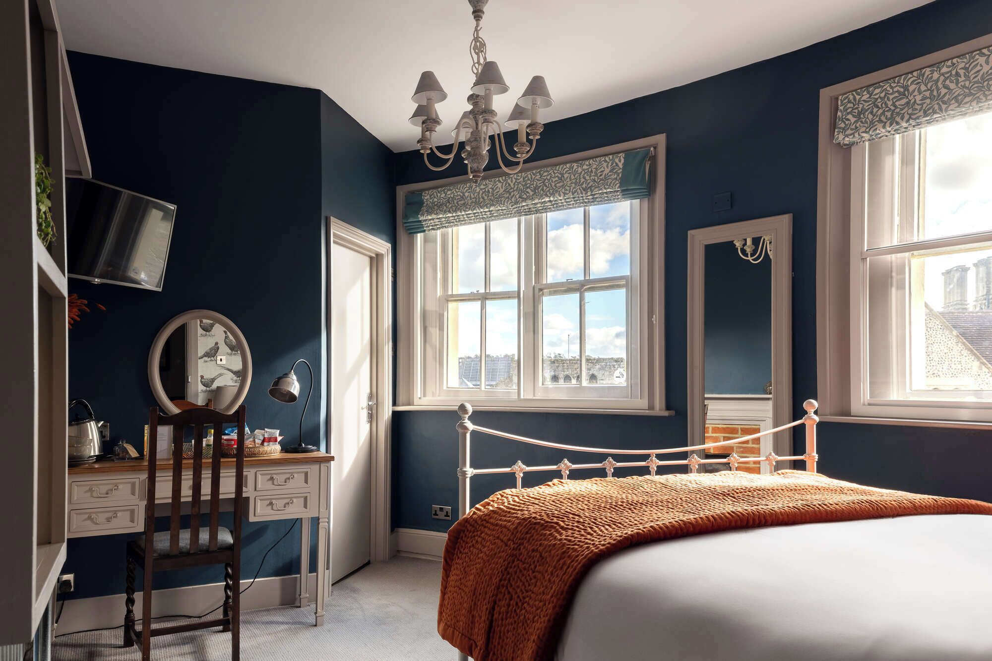 A bedroom with a bed, a desk, a chair, a mirror, and a window with a view of buildings.