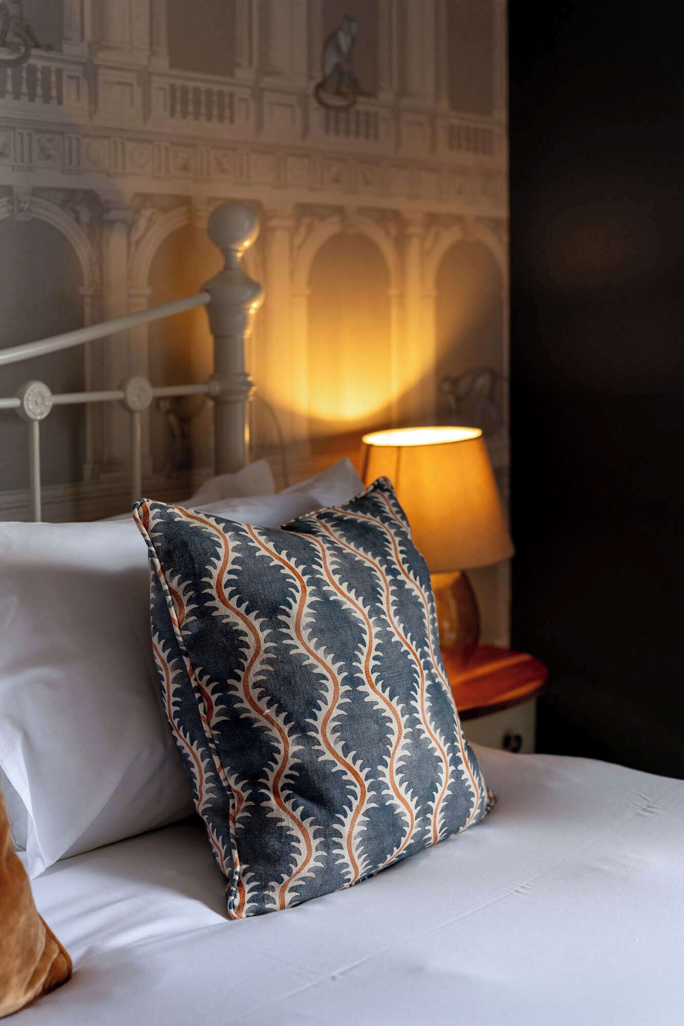 A bed with a patterned pillow, a nightstand with a lamp, and a wall with architectural details.