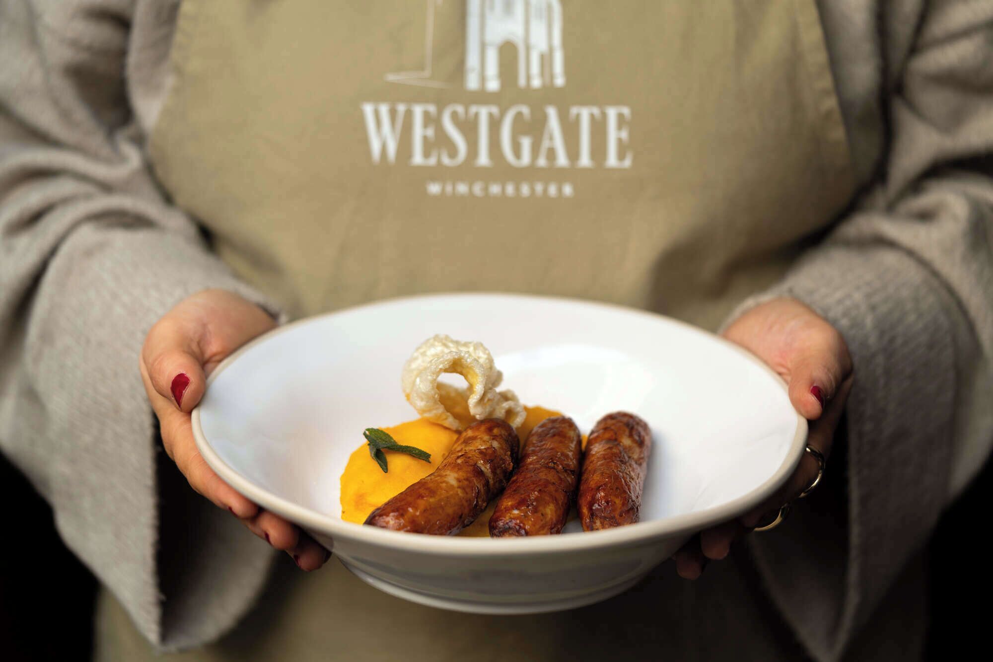 Une personne tenant un bol rempli de saucisses et un morceau d’aliment jaune, et portant un tablier sur lequel est écrit le mot « WESTGATE ».