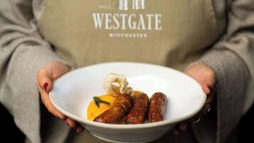 Une personne tenant un bol rempli de saucisses et un morceau d’aliment jaune, et portant un tablier sur lequel est écrit le mot « WESTGATE ».