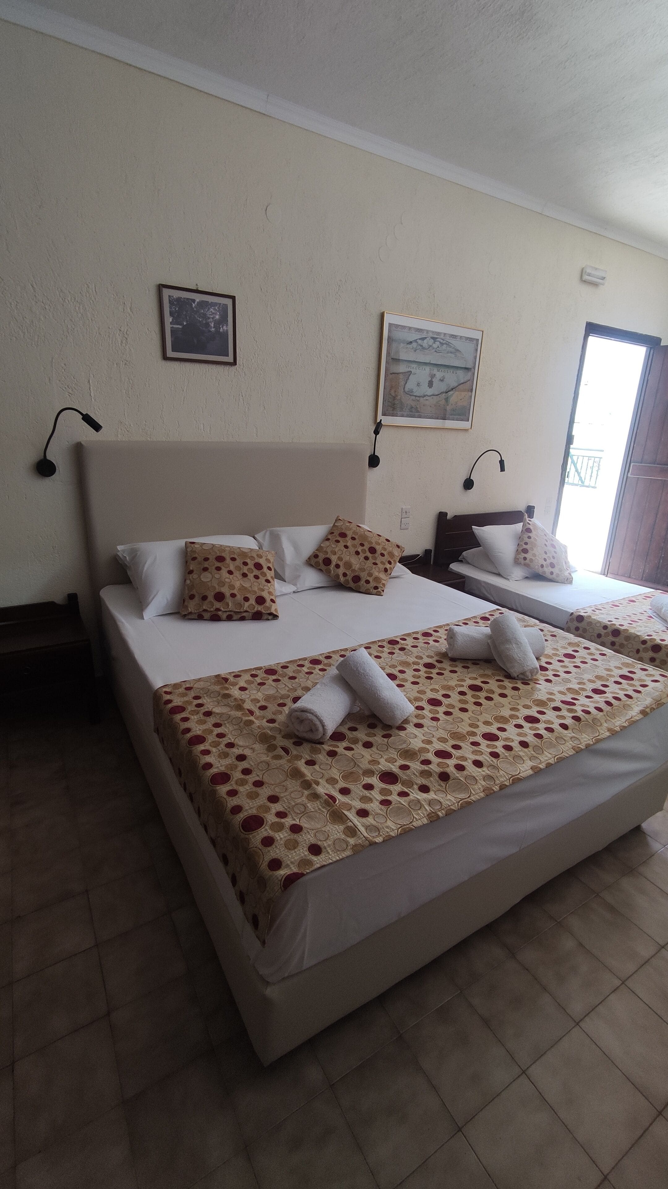 A hotel room with two beds, each with patterned bedding and pillows. There are wall-mounted lamps and a framed picture on the wall.