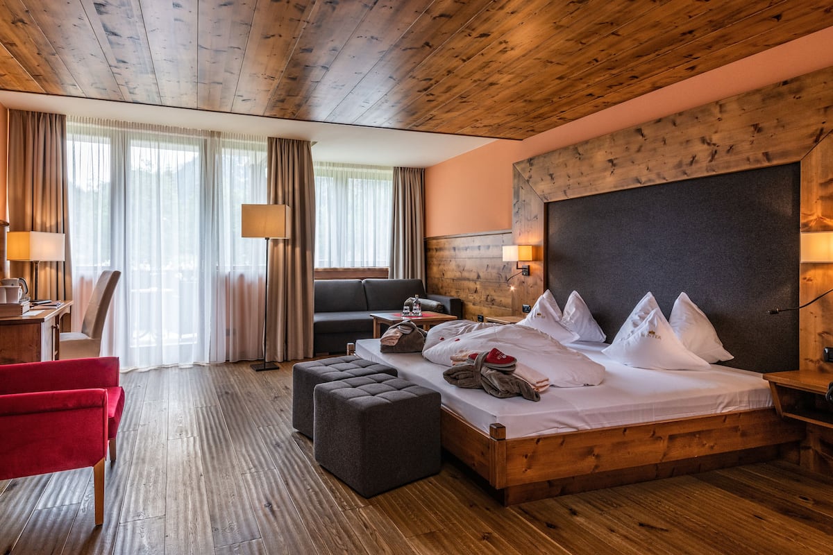 A hotel room with a wooden ceiling, a bed with white linens and multiple pillows, a red armchair, a wooden desk with a lamp, and a large window with curtains.