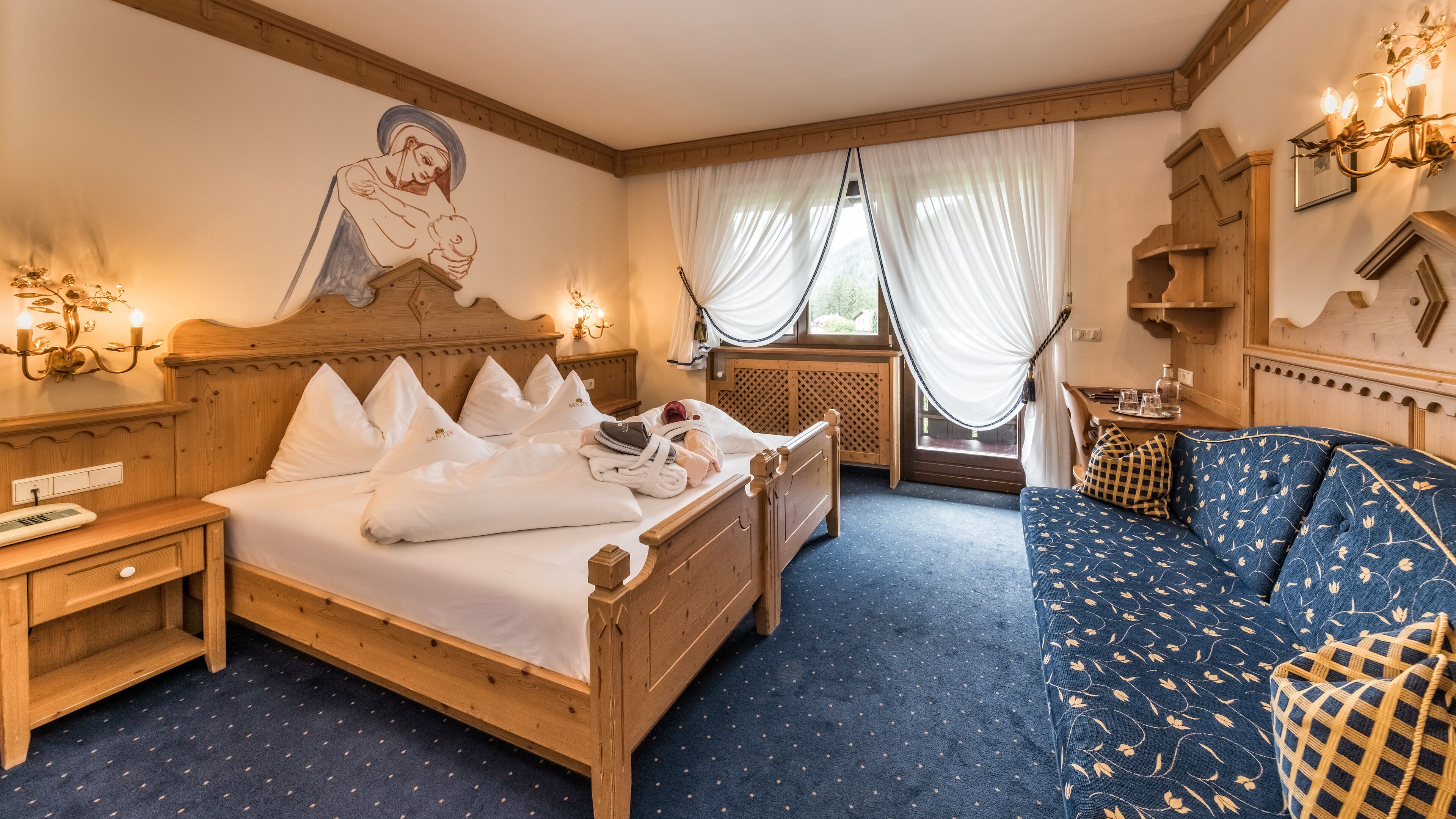 A hotel room with a wooden bed, a blue sofa, a wall-mounted artwork, and a window with sheer curtains.