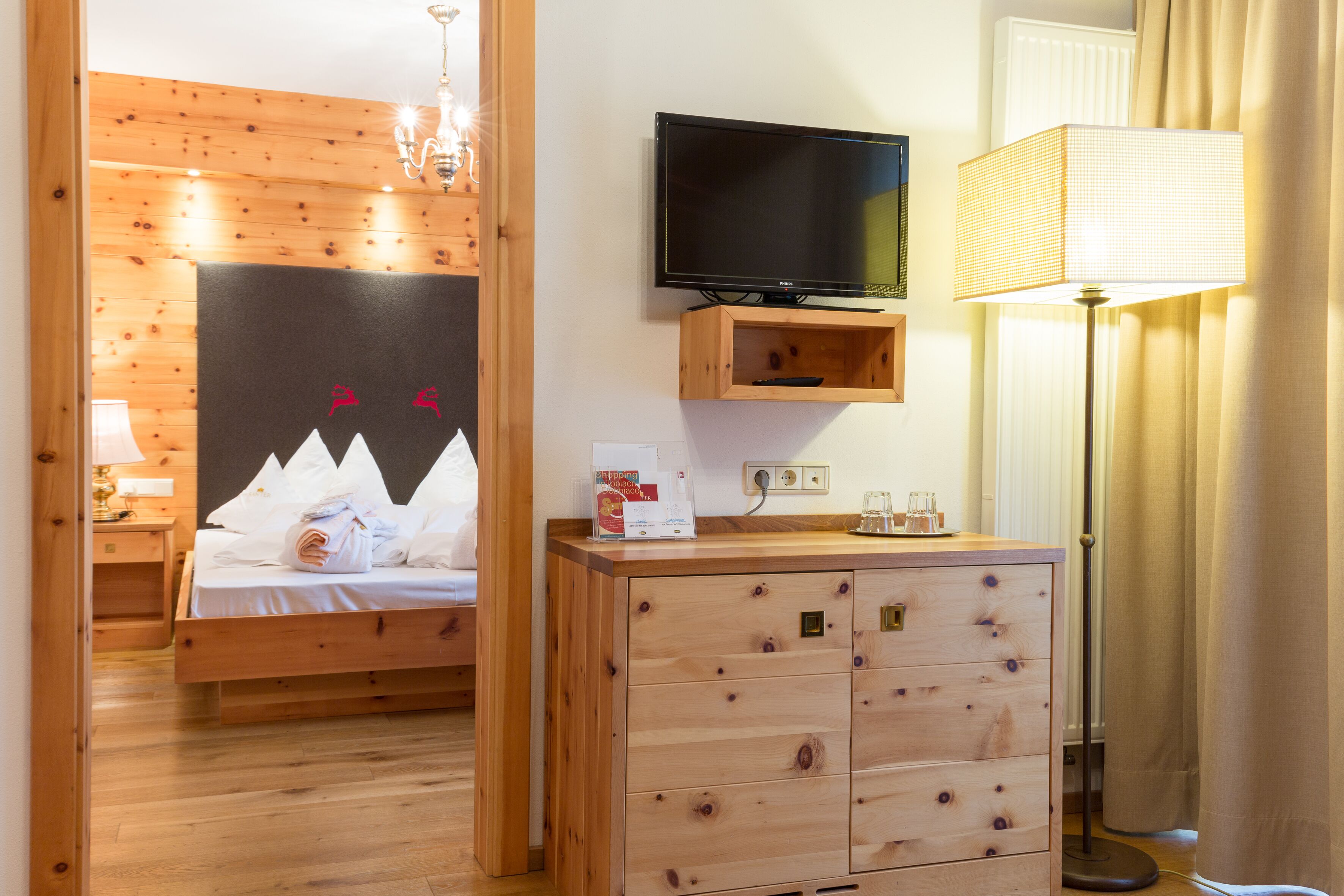 A wooden bedroom with a bed, a TV mounted on a wooden shelf, a bedside table, and a lamp.