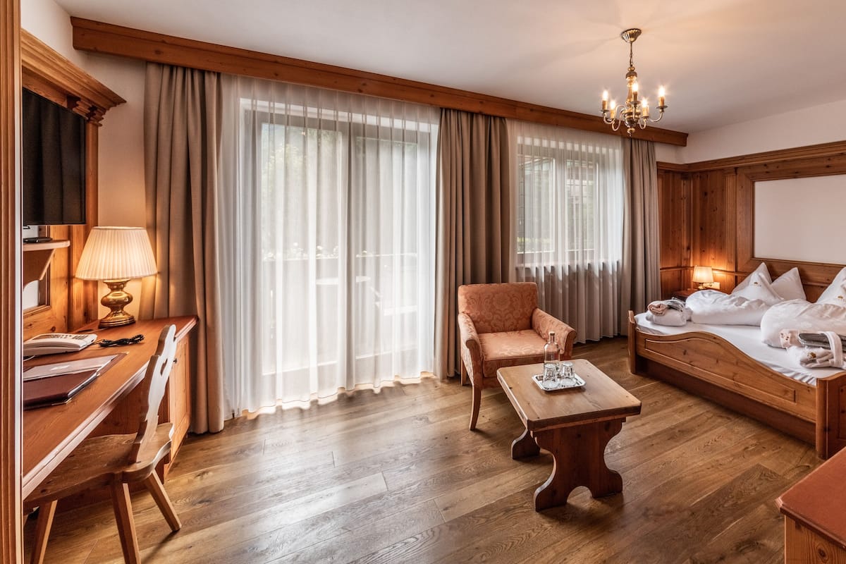A hotel room with a wooden bed, a wooden desk, a chair, a coffee table, and a large window with sheer curtains.