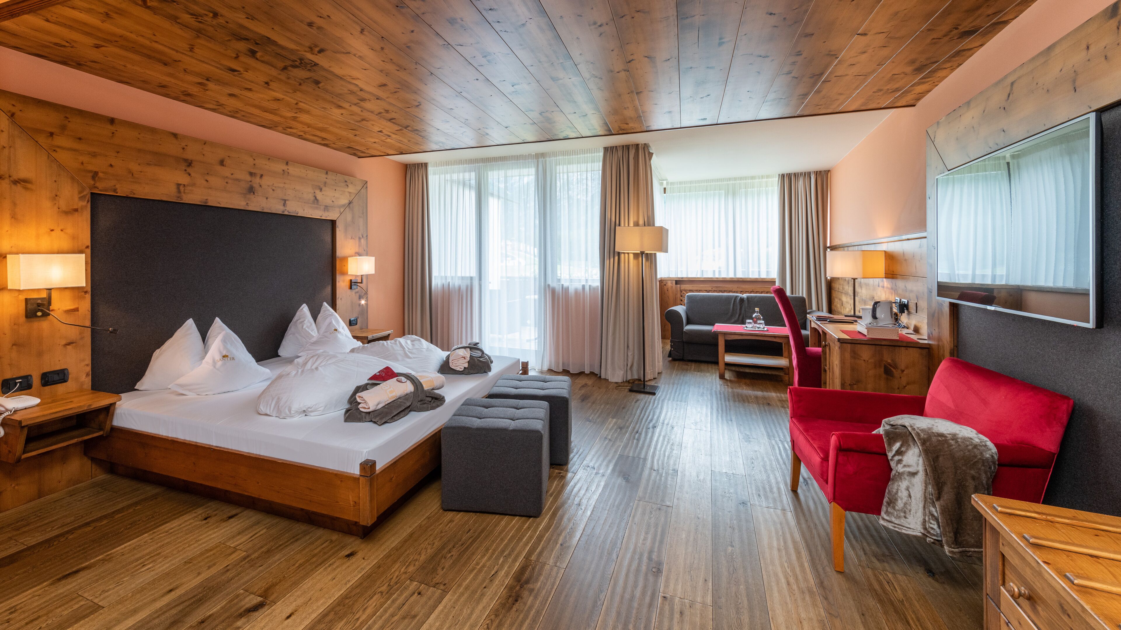 Une chambre d’hôtel avec un plafond en bois, un grand lit recouvert de draps blancs, un fauteuil rouge, un bureau en bois et un téléviseur fixé au mur.