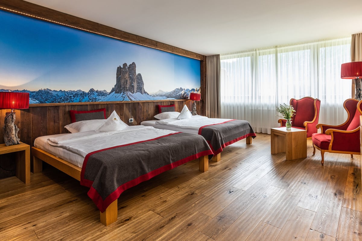 A hotel room with two beds, wooden flooring, a red armchair, a wooden desk, a red lamp, and a painting of a mountain landscape.