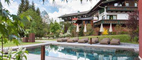 A pool area with a wooden deck, lounge chairs, and a building with a restaurant bar in the background.