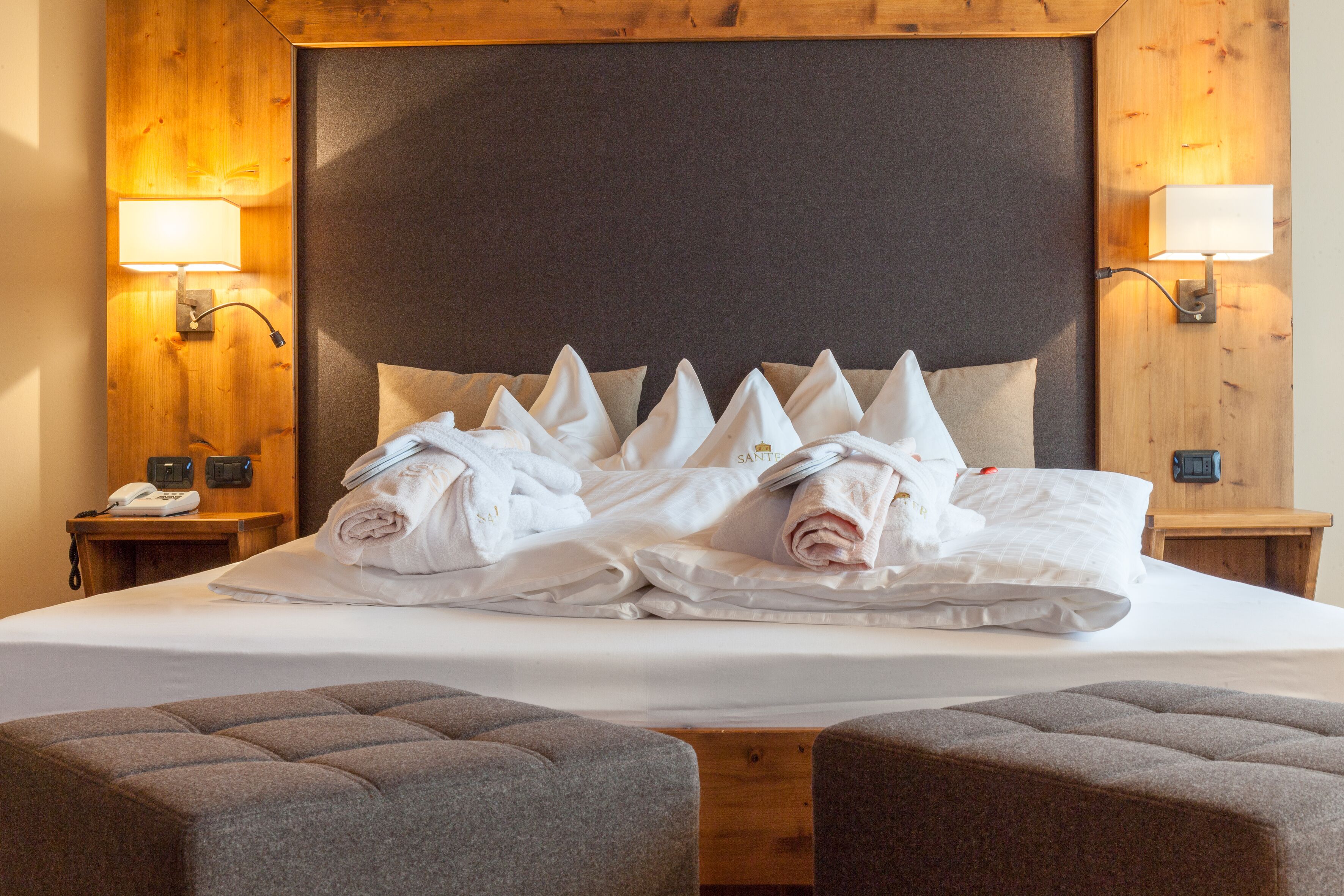 A neatly made bed with towels and pillows, wooden headboard, two bedside lamps, and a small table with a phone and a clock.