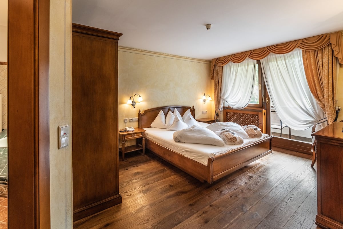 A hotel room with a wooden bed, a nightstand with a lamp, a bathroom with a towel rack, and a view of the outdoors through the window.