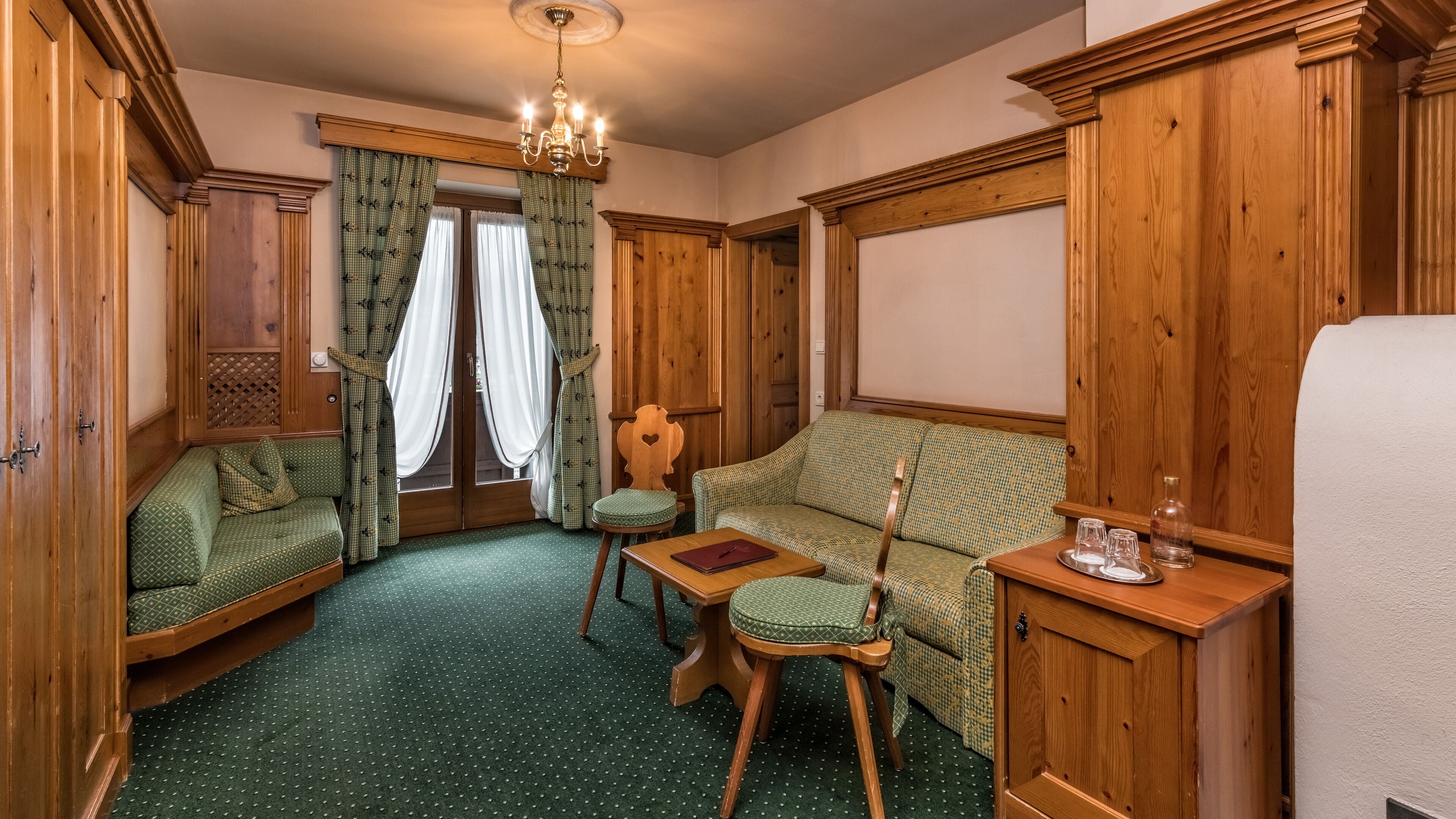 Una habitación acogedora con un sofá verde estampado, una silla de madera, una mesita con vasos y un armario de madera.