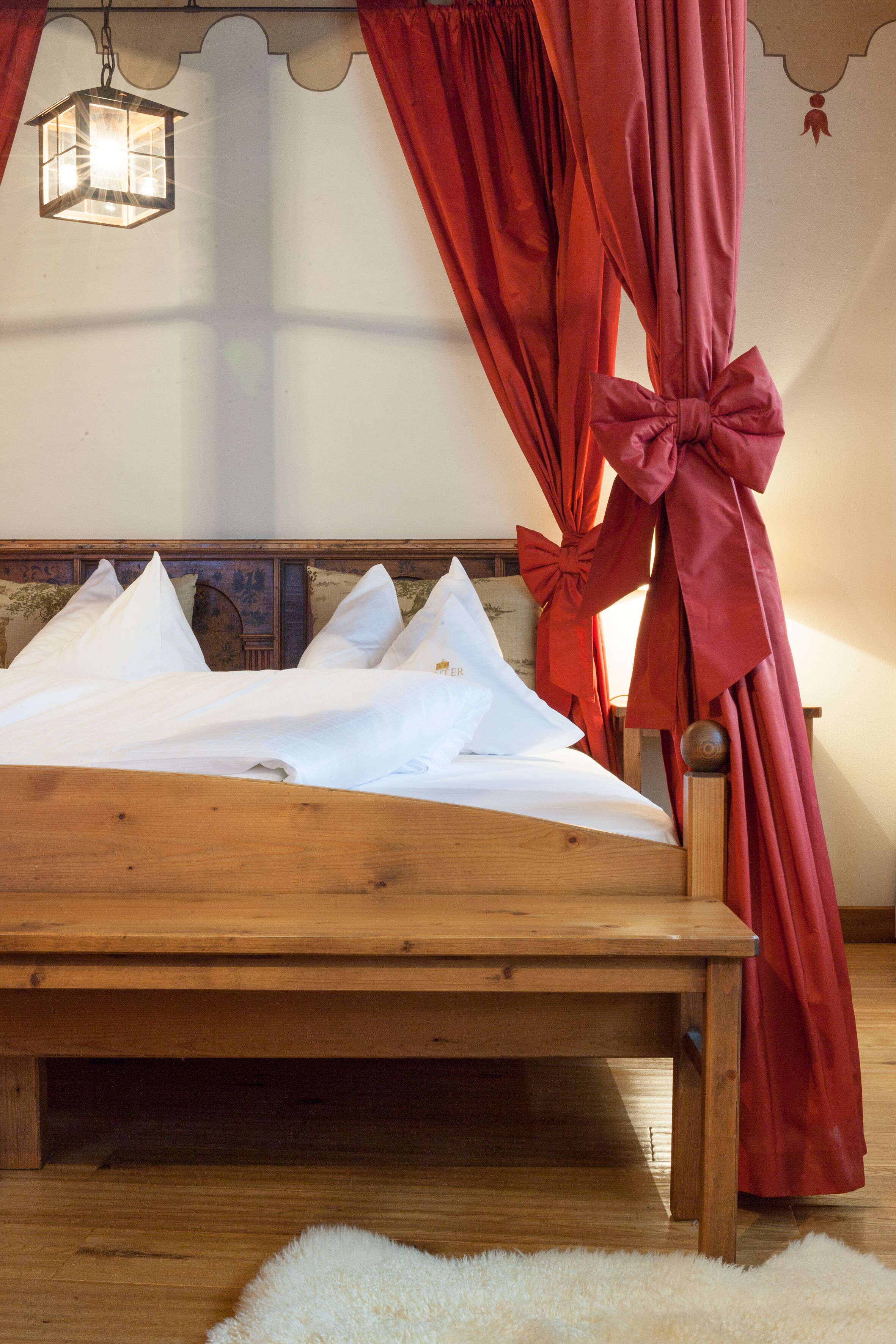 A neatly made bed with white linens, a wooden headboard, and red curtains with bows.