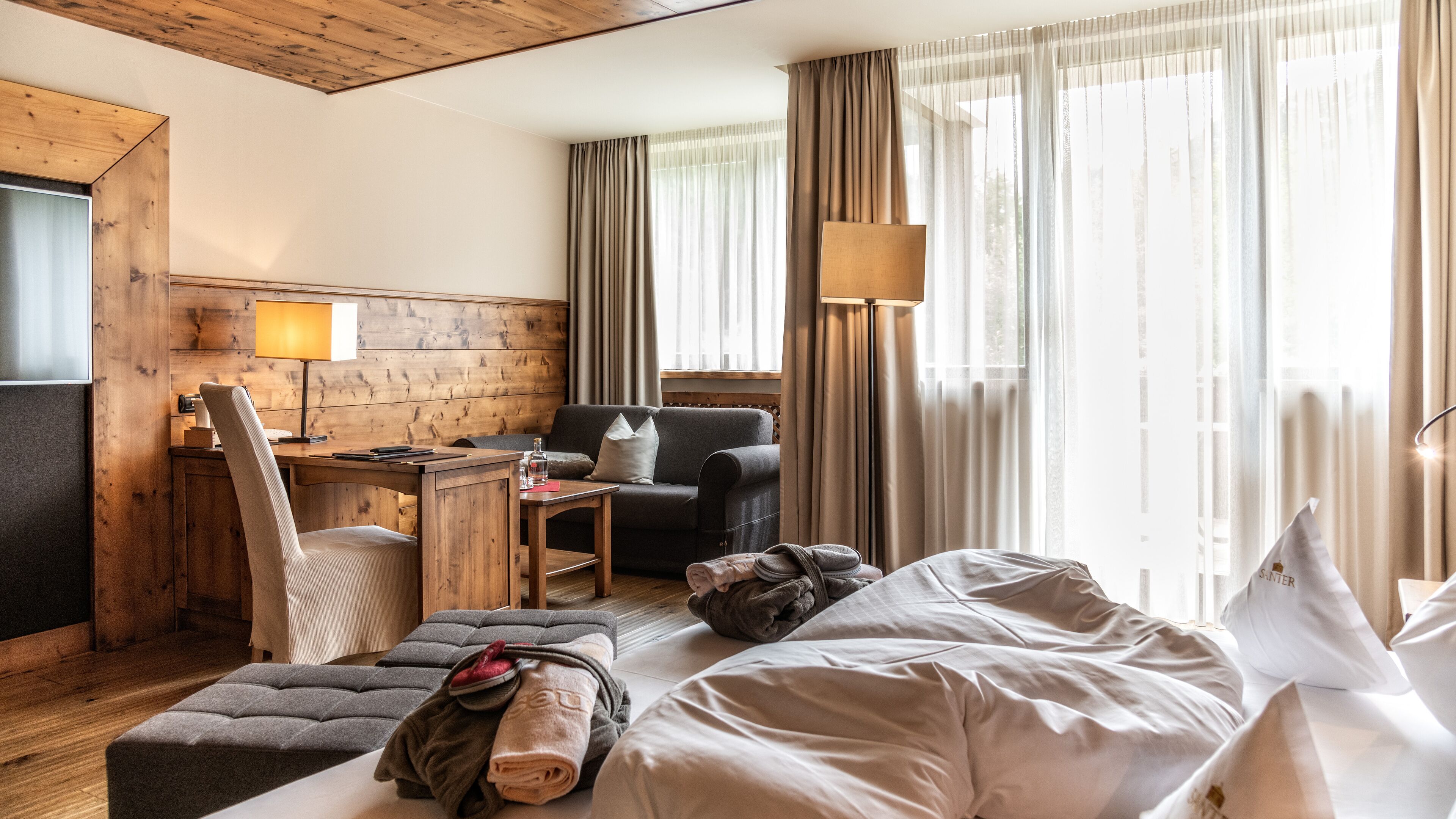 Camera d'albergo con un poggiatesta in legno, una scrivania con una lampada, un divano, un letto con asciugamani e ampie finestre con tende.