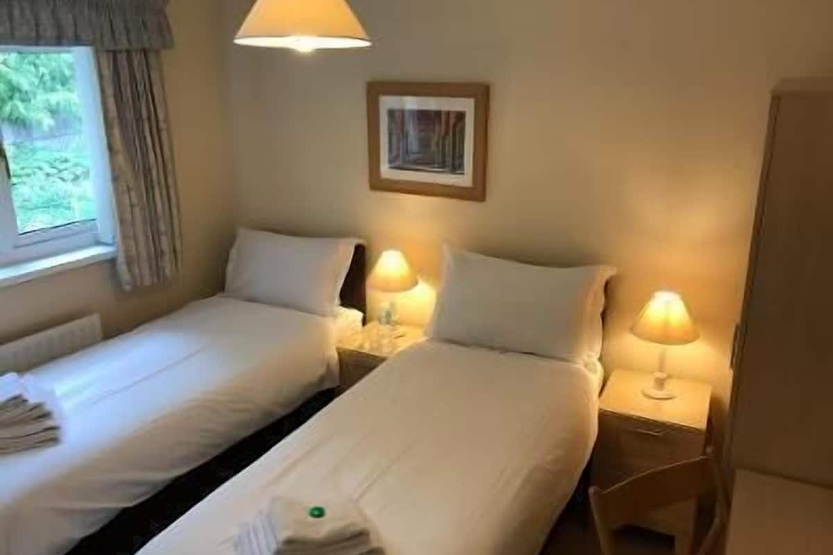 Ein Hotelzimmer mit zwei Einzelbetten, Nachttischlampen, einem Fenster mit Vorhängen, einem gerahmten Bild an der Wand und einem Heizkörper unter dem Fenster.