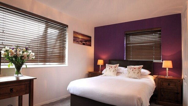 A bedroom with a large bed, two bedside tables with lamps, a nightstand, and a vase of flowers on a desk. - Abbey Bed and Breakfast (Londonderry)
