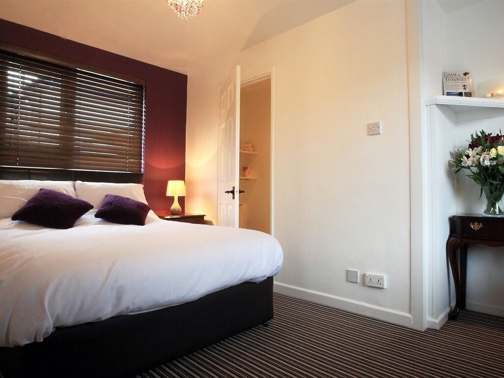 A hotel room with a large bed, a bedside table with a lamp, a wardrobe, and a shelf with books and flowers.