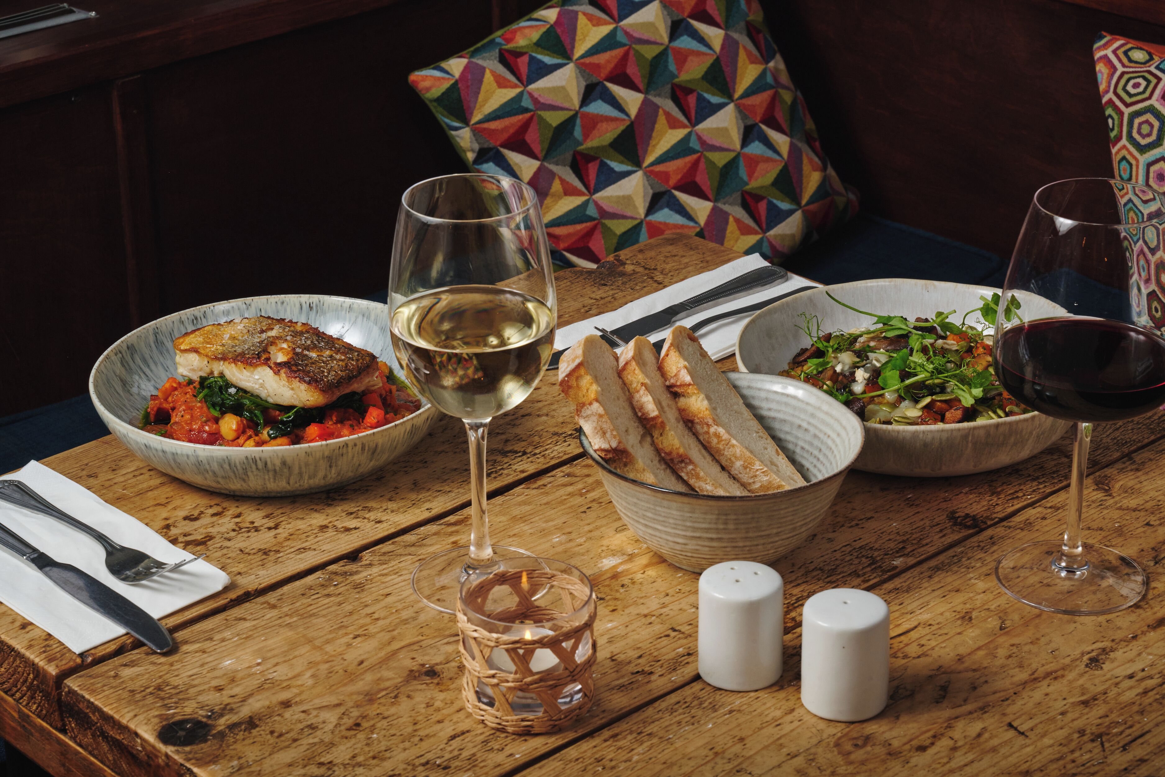 A wooden table with a meal consisting of fish, bread, and salad, accompanied by wine glasses.