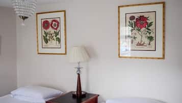 Habitación de hotel con dos camas, una mesita de noche, una lámpara e ilustraciones botánicas enmarcadas en la pared.