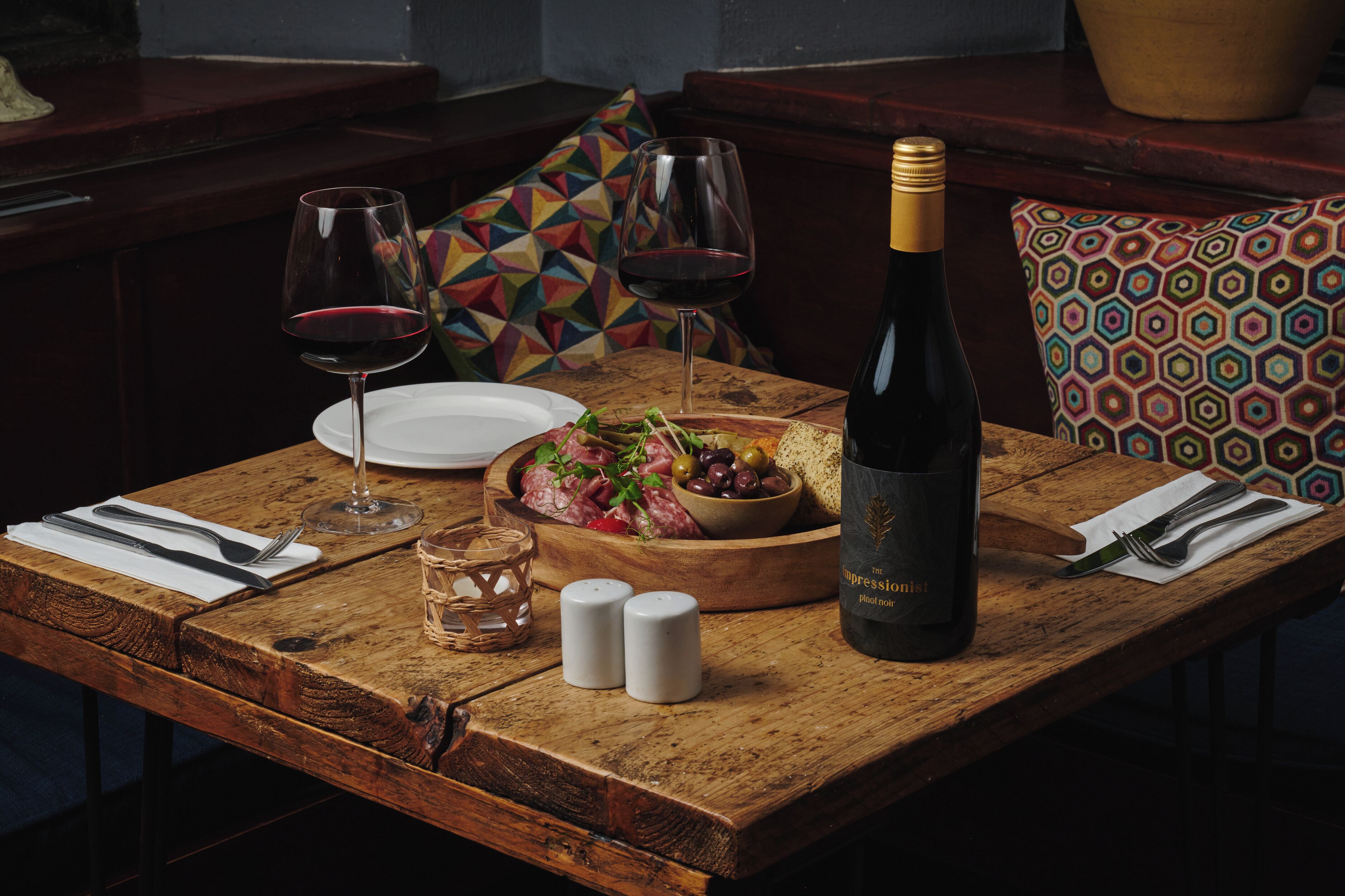 A wooden dining table set with a bottle of wine, glasses, a bowl of salad, and a plate.