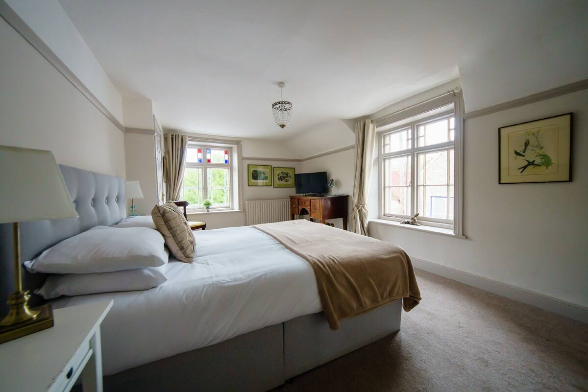 A bedroom with a large bed, a bedside table with a lamp, a dresser, and a window with curtains.