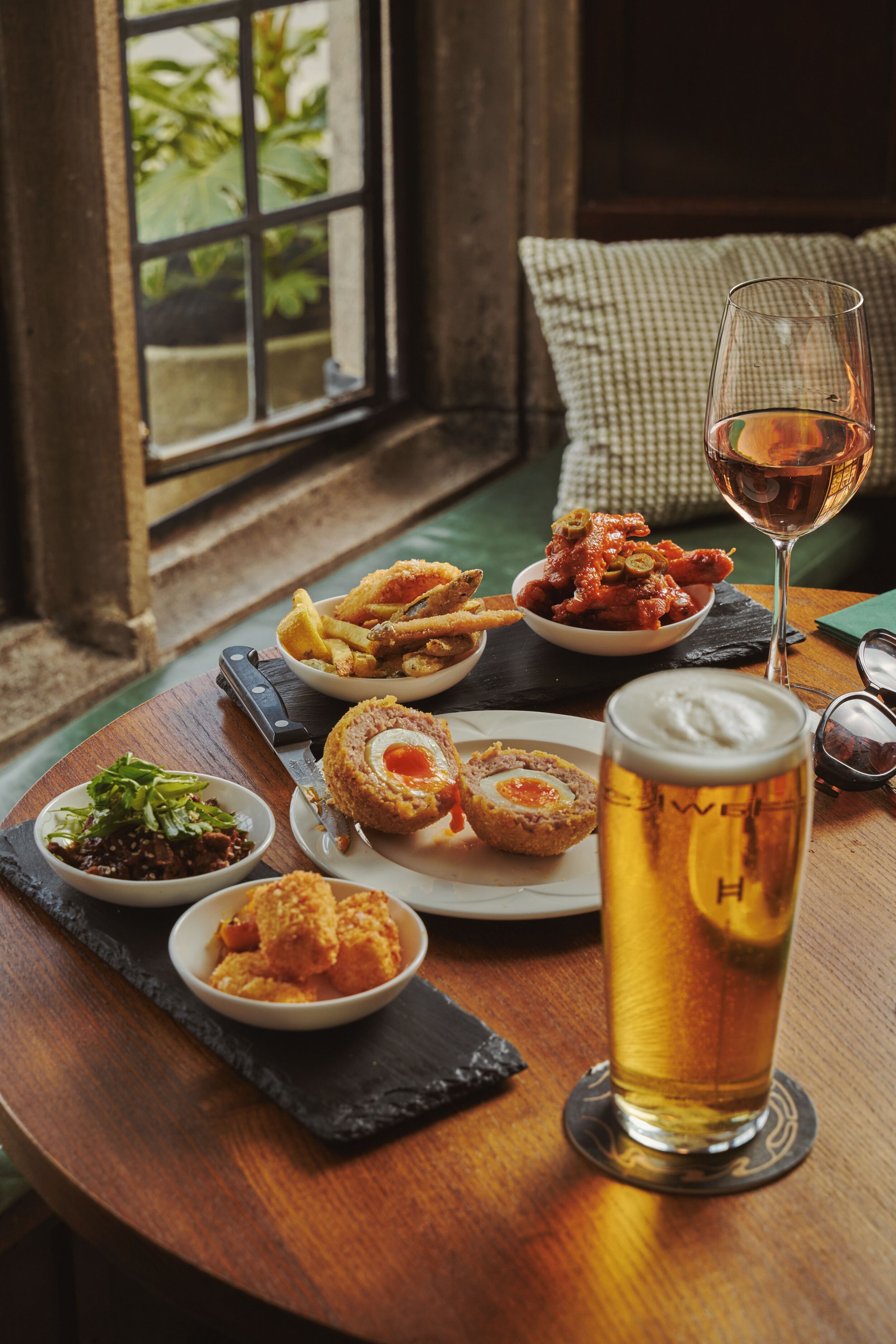 A wooden table with a variety of dishes, a glass of beer, and a window view.