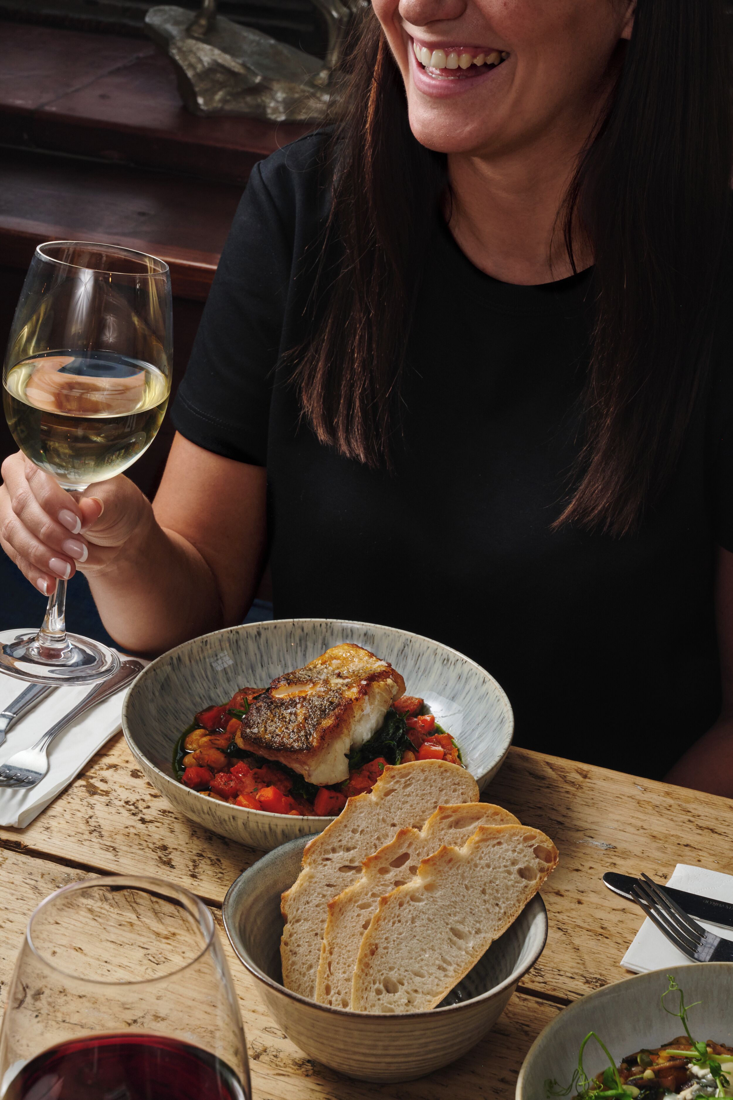 A person seated at a wooden table with a bowl of food, a glass of white wine, and a plate of bread.
