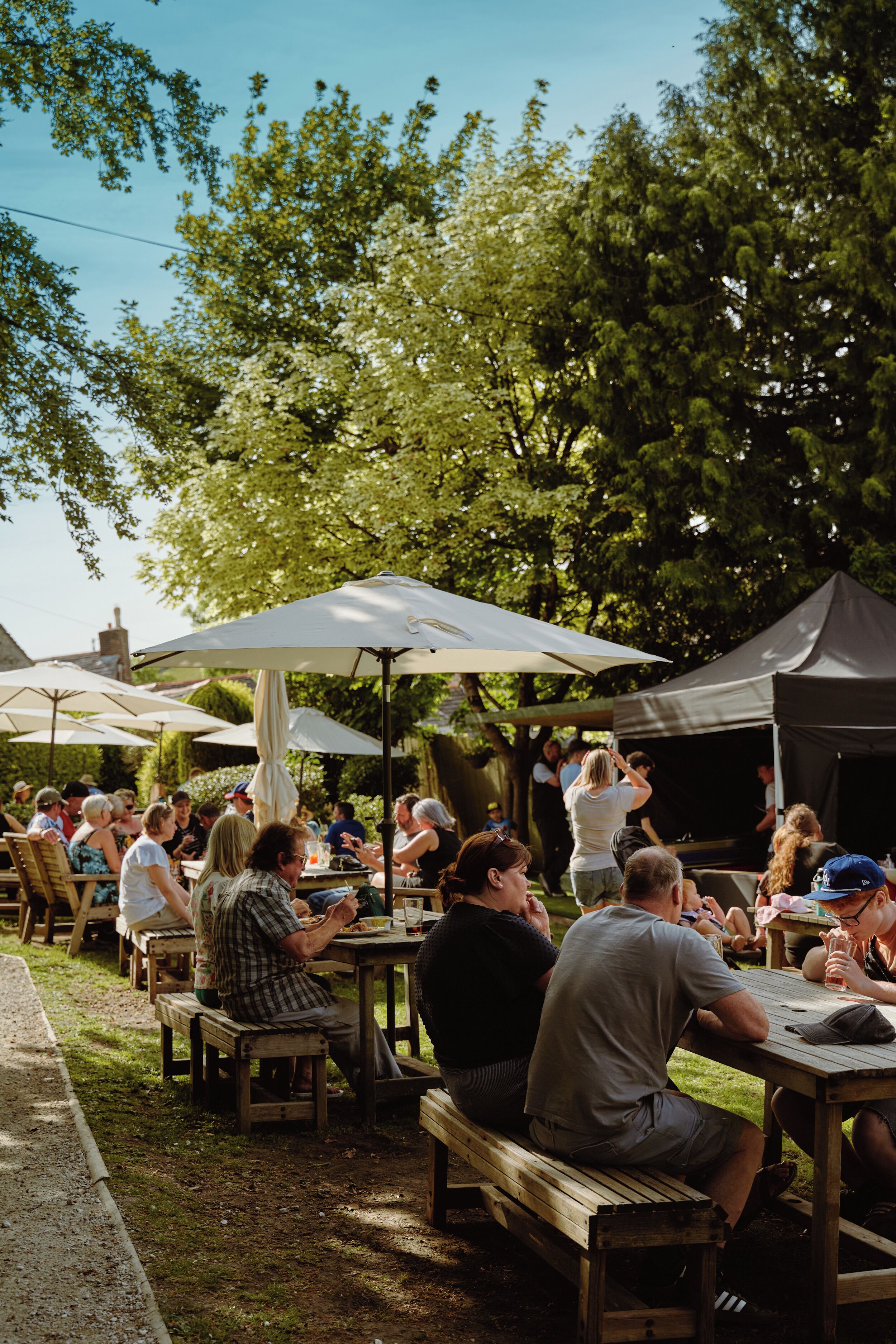 An outdoor dining area with wooden tables and benches, surrounded by trees and covered by umbrellas.