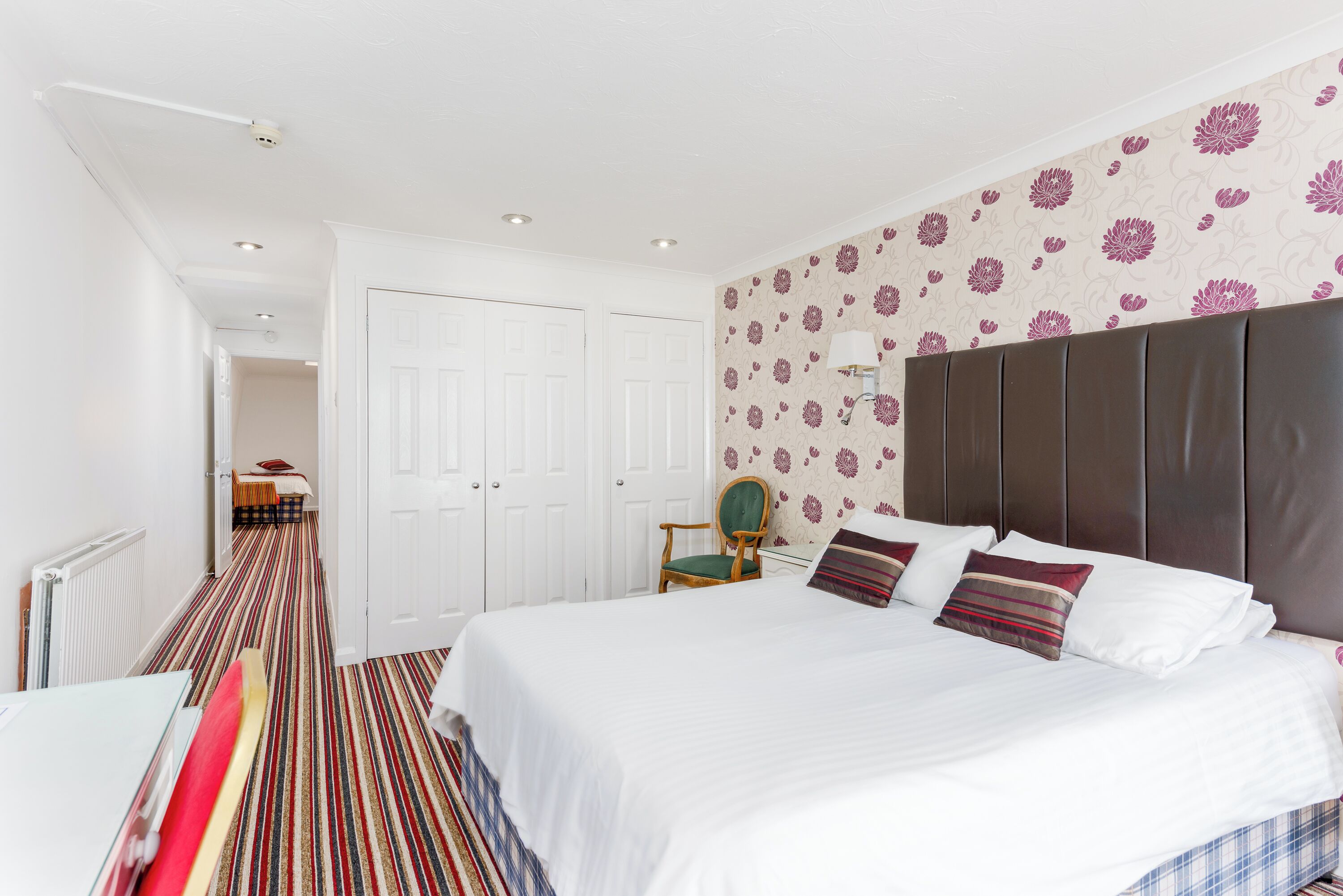 A bedroom with a large bed, a chair, a wardrobe, and a patterned wall.