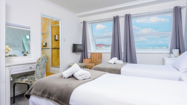 A hotel room with two beds, a desk with a chair, a TV, and a window with curtains. - Barrowfield Hotel (Newquay)