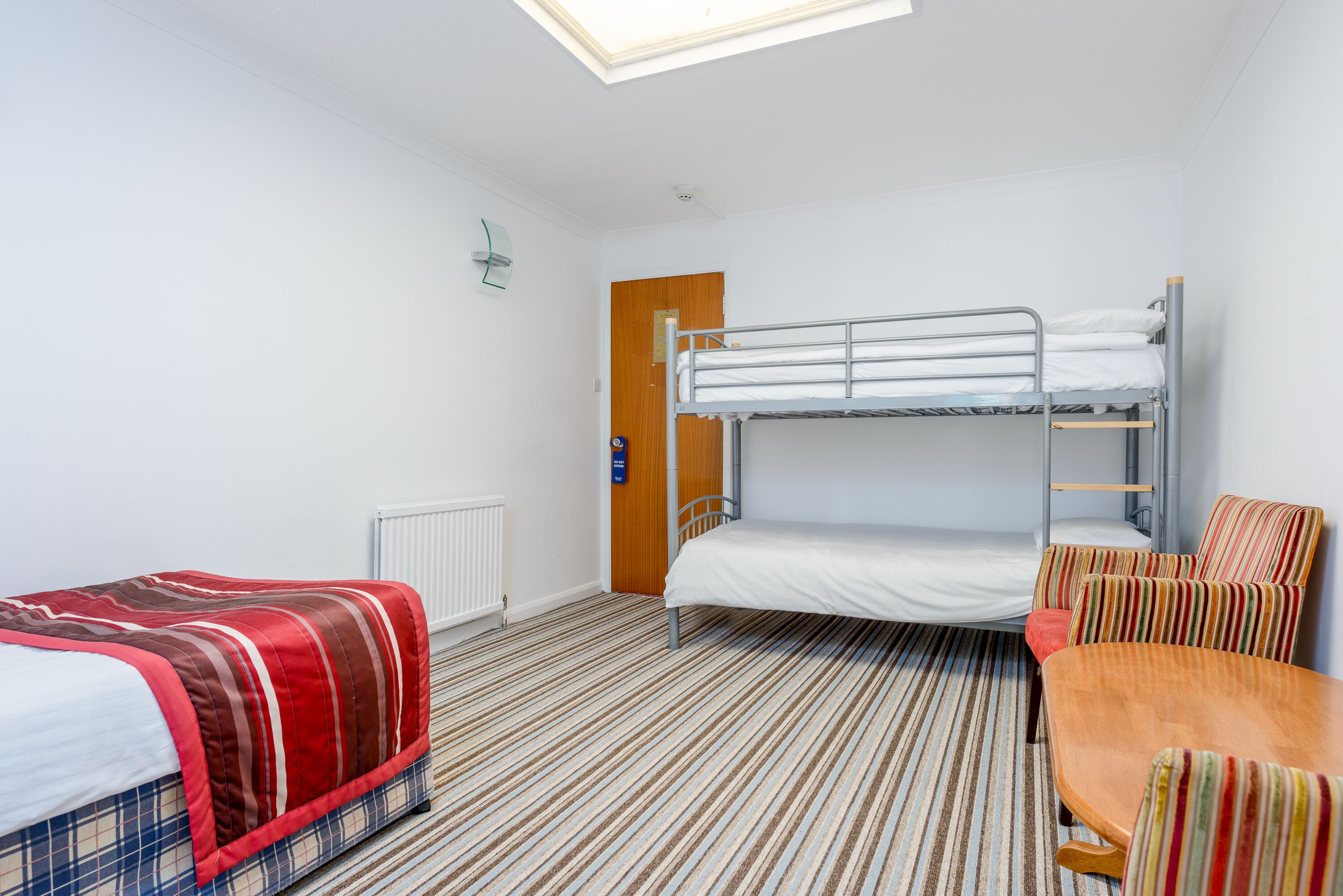 A room with a bunk bed, a single bed with a striped blanket, a wooden table, and a striped chair.