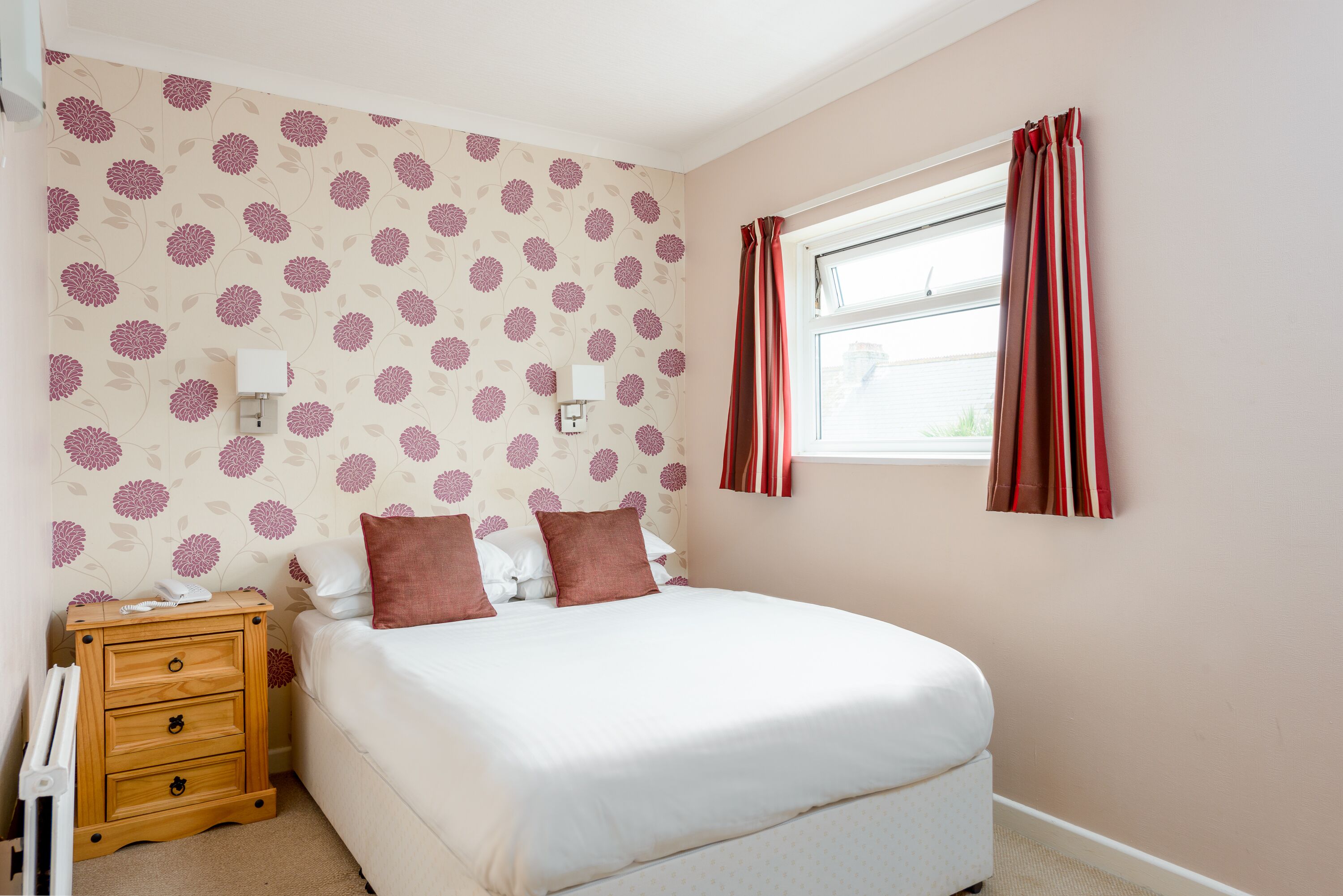 A bedroom with a bed, a nightstand, a wall-mounted light, and a window with curtains.