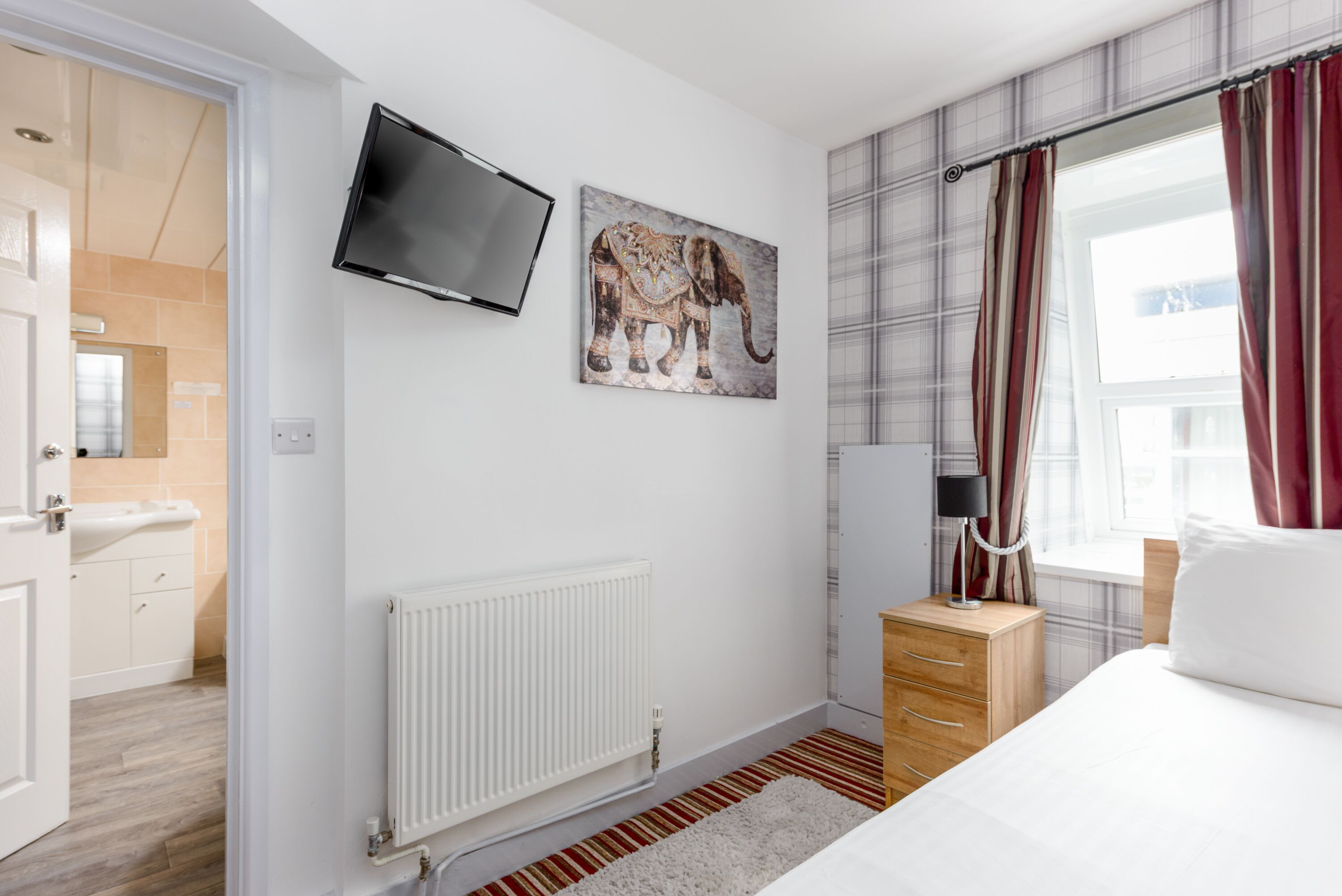 A hotel room with a bed, a TV mounted on the wall, a radiator, a wooden nightstand, and a window with curtains.