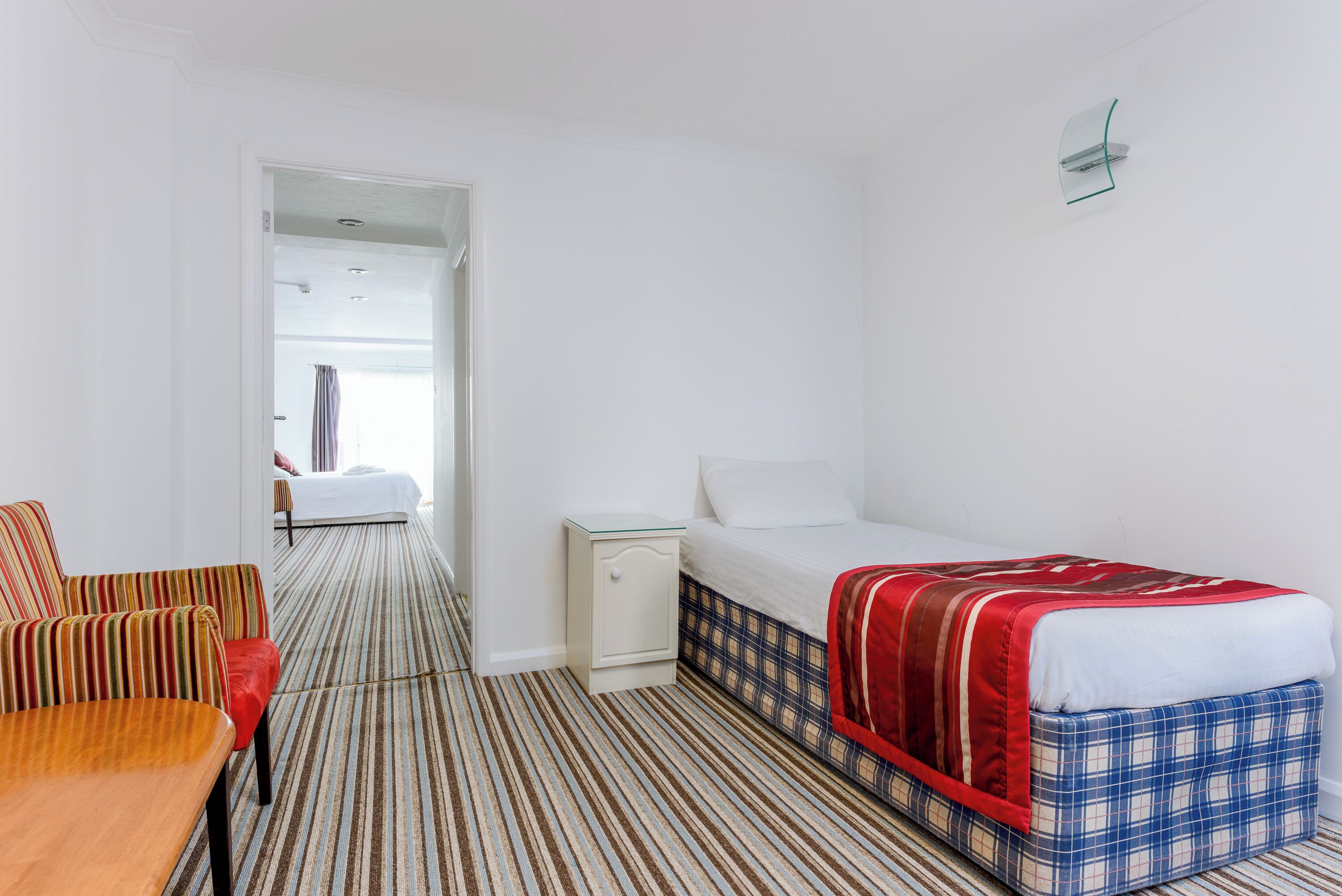 Une chambre d’hôtel avec une moquette à rayures, un lit recouvert d’une couverture à carreaux, une petite table de chevet et une chaise avec un coussin à rayures.