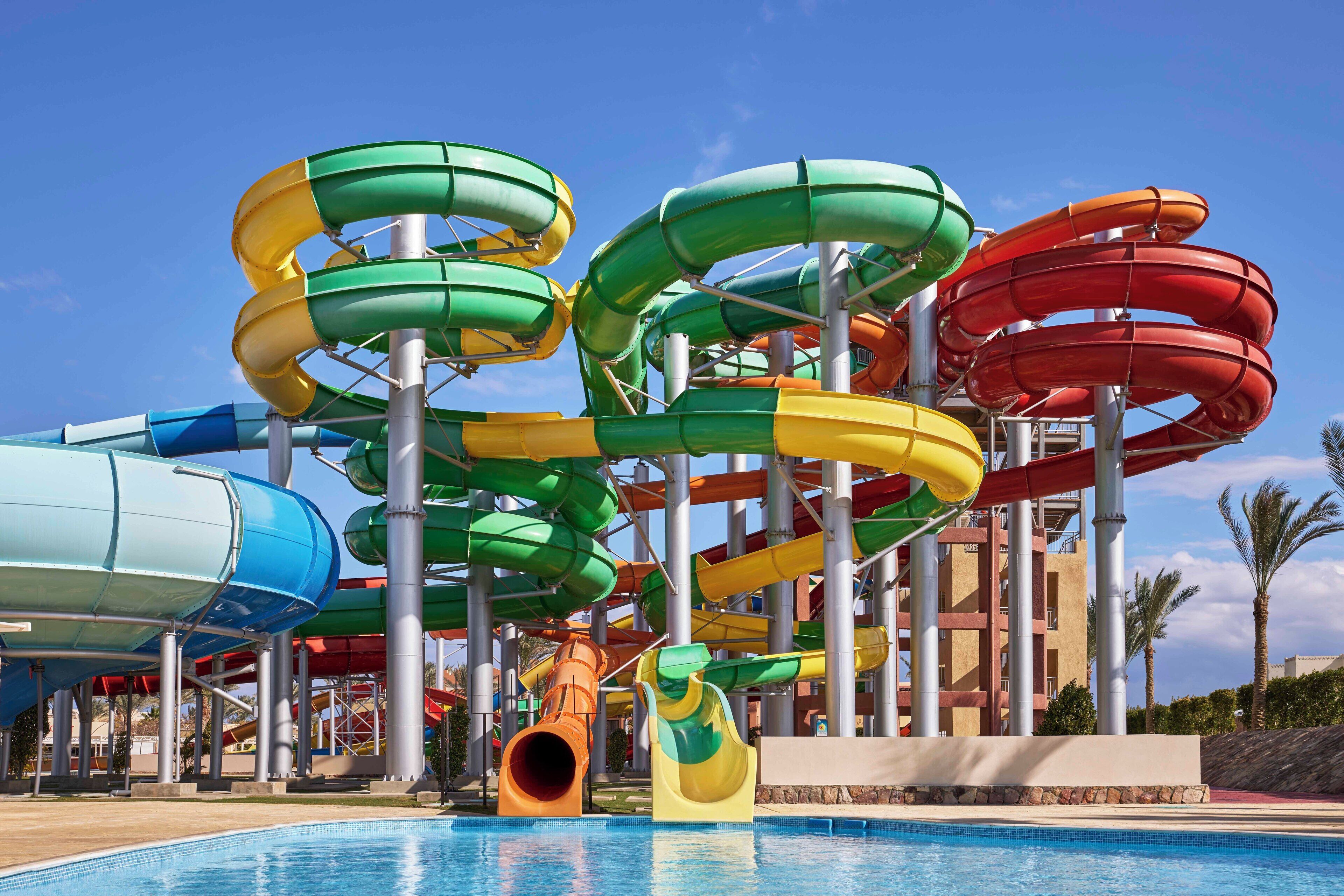 Parque acuático con múltiples toboganes de agua de varios colores, una piscina y una estructura de edificio al fondo.