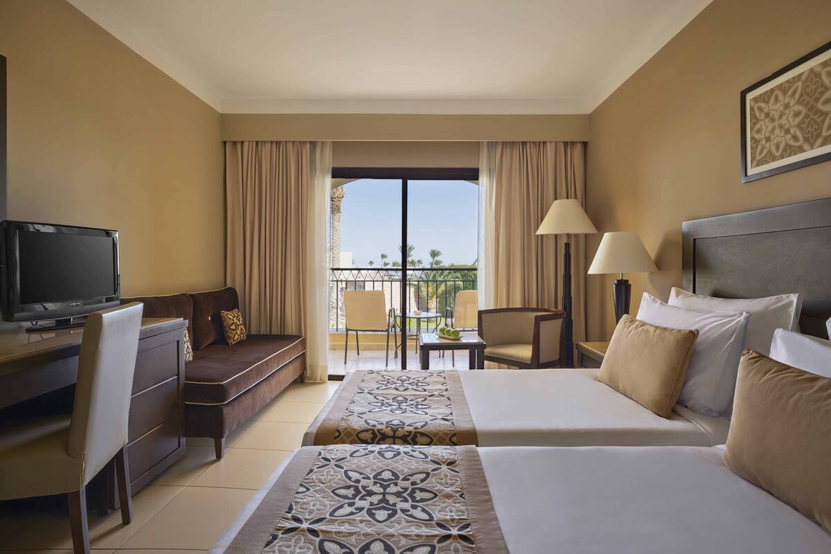 Quarto de hotel com cama, escrivaninha, televisão, sofá e varanda com cadeiras.