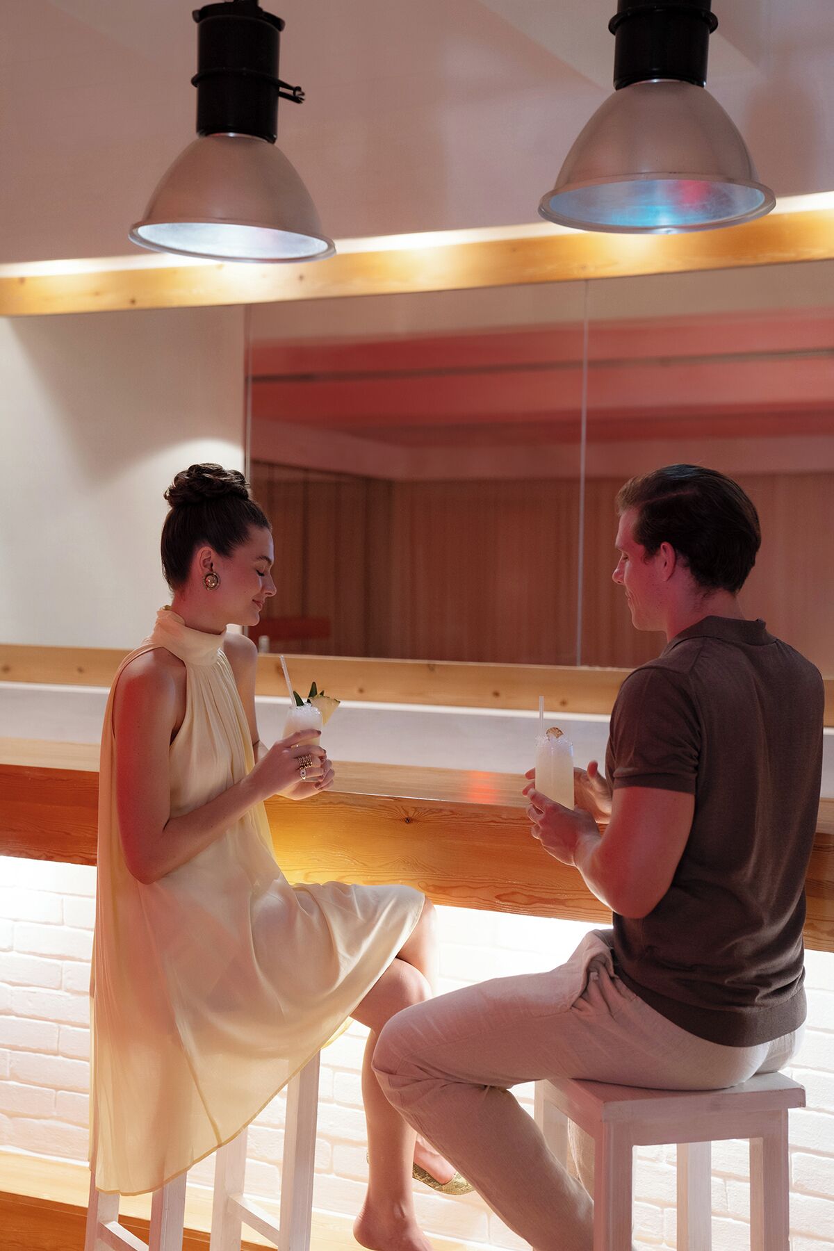Two people sitting on white stools in a room with wooden flooring and a large mirror.