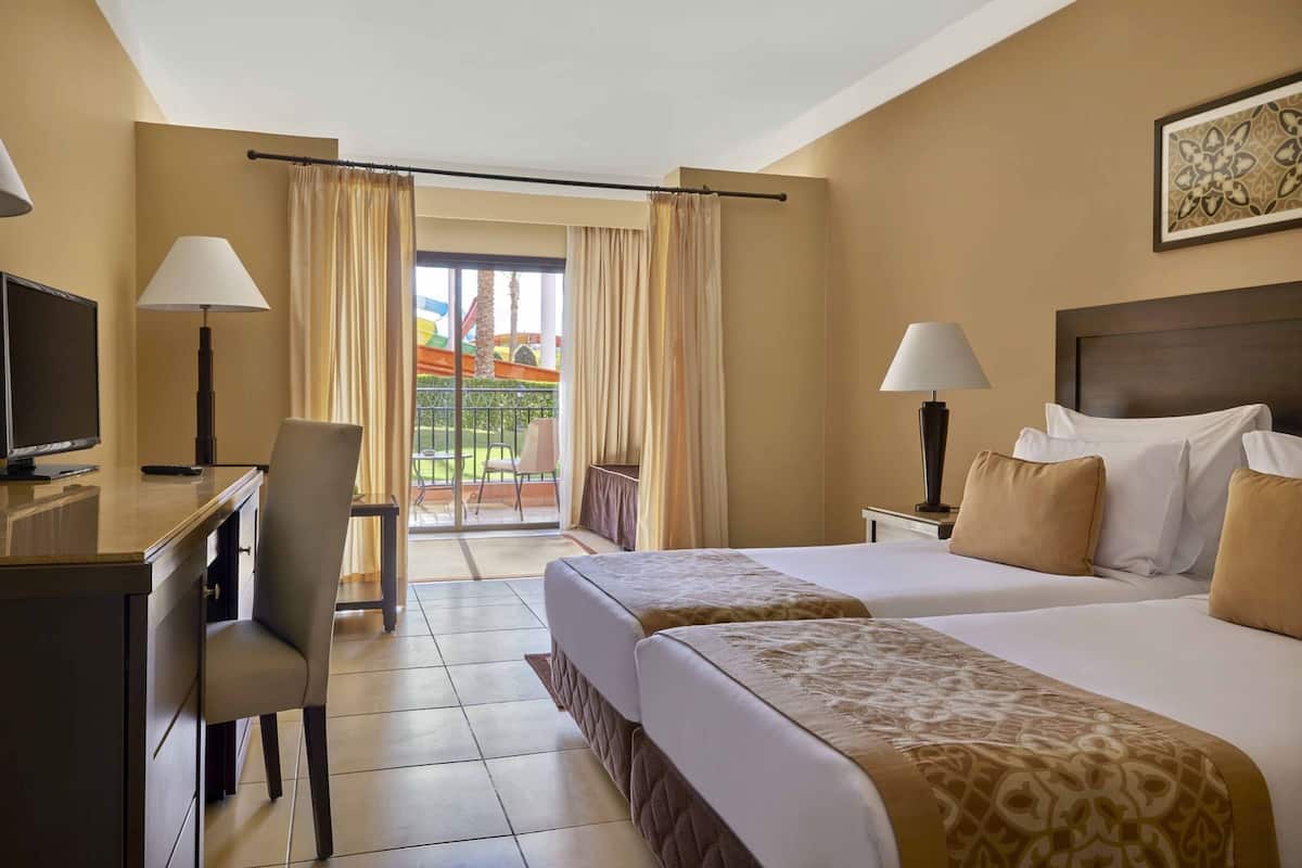 Quarto de hotel com cama, escrivaninha, cadeira, televisão e varanda com cortinas.