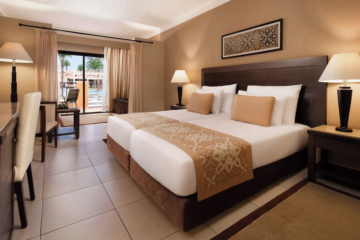 Quarto de hotel com uma cama grande, duas mesas de cabeceira com luminárias, uma escrivaninha com cadeira e vista para a área da piscina.