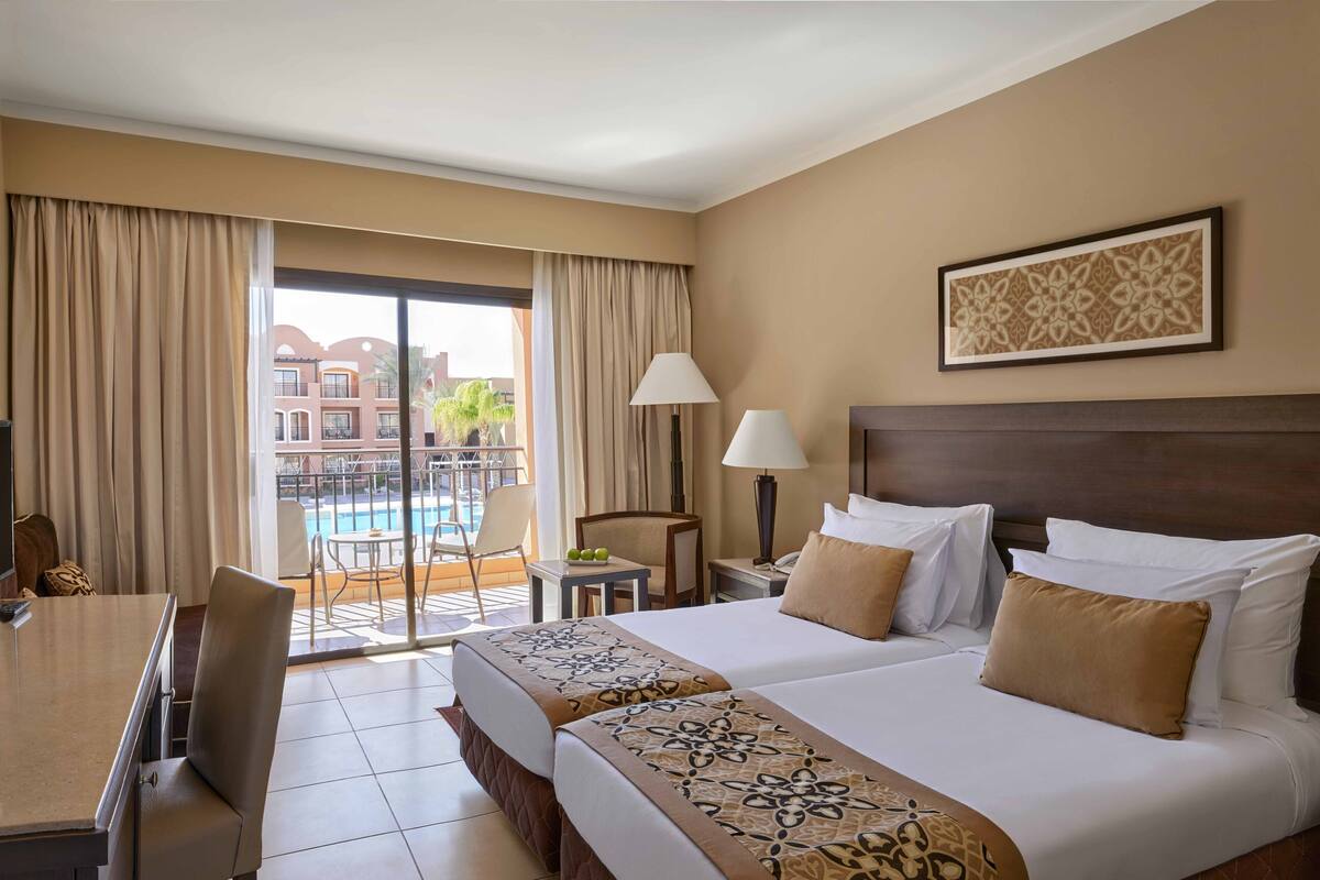 Ein Hotelzimmer mit einem Bett, einem Schreibtisch, einem Sessel, einer Lampe, einem Tisch mit Obst und Blick auf Gebäude sowie einen Pool.