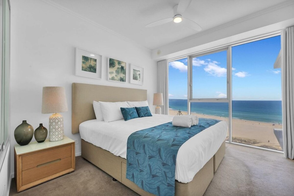 A bedroom with a large window, a bed, a nightstand, and wall art.