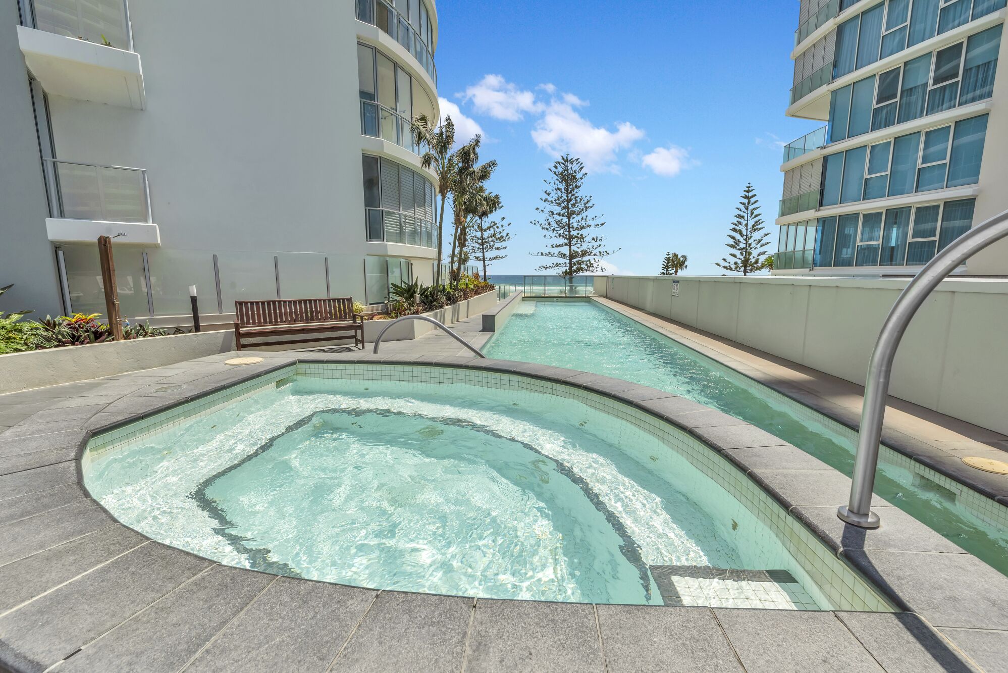 A swimming pool area with a curved pool, a bench, and a view of the ocean.