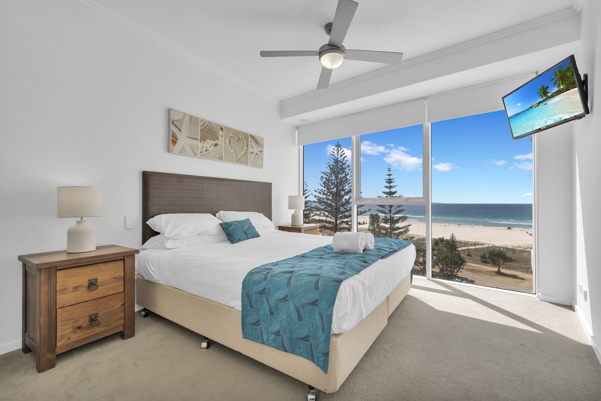 A bedroom with a bed, bedside table, lamp, artwork, ceiling fan, and a TV.