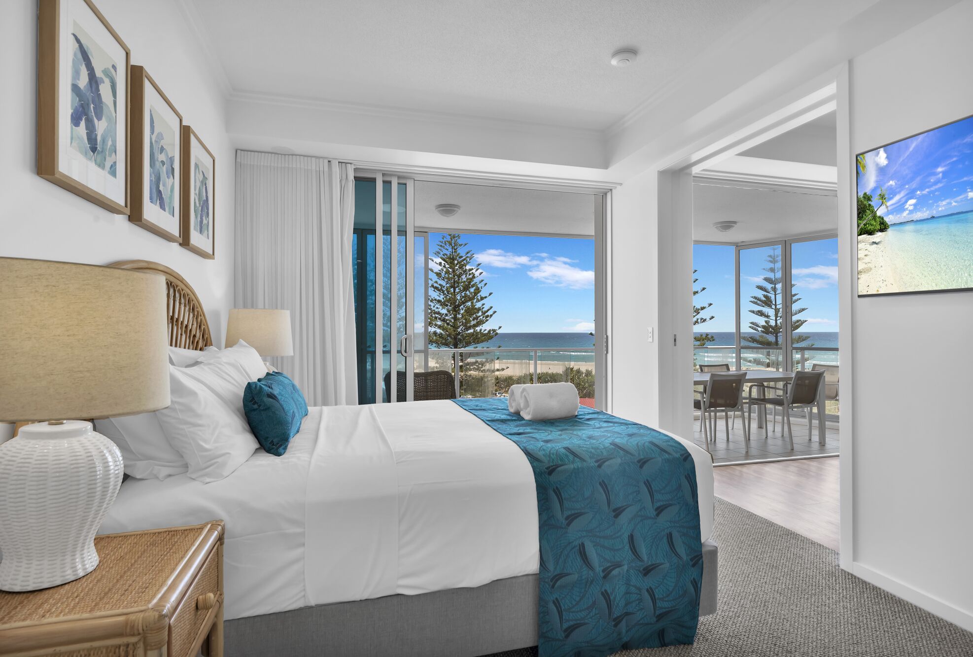 Une chambre d’hôtel comprenant un lit, une table de chevet, une lampe, une fenêtre donnant sur la plage et un balcon avec une table et des chaises.