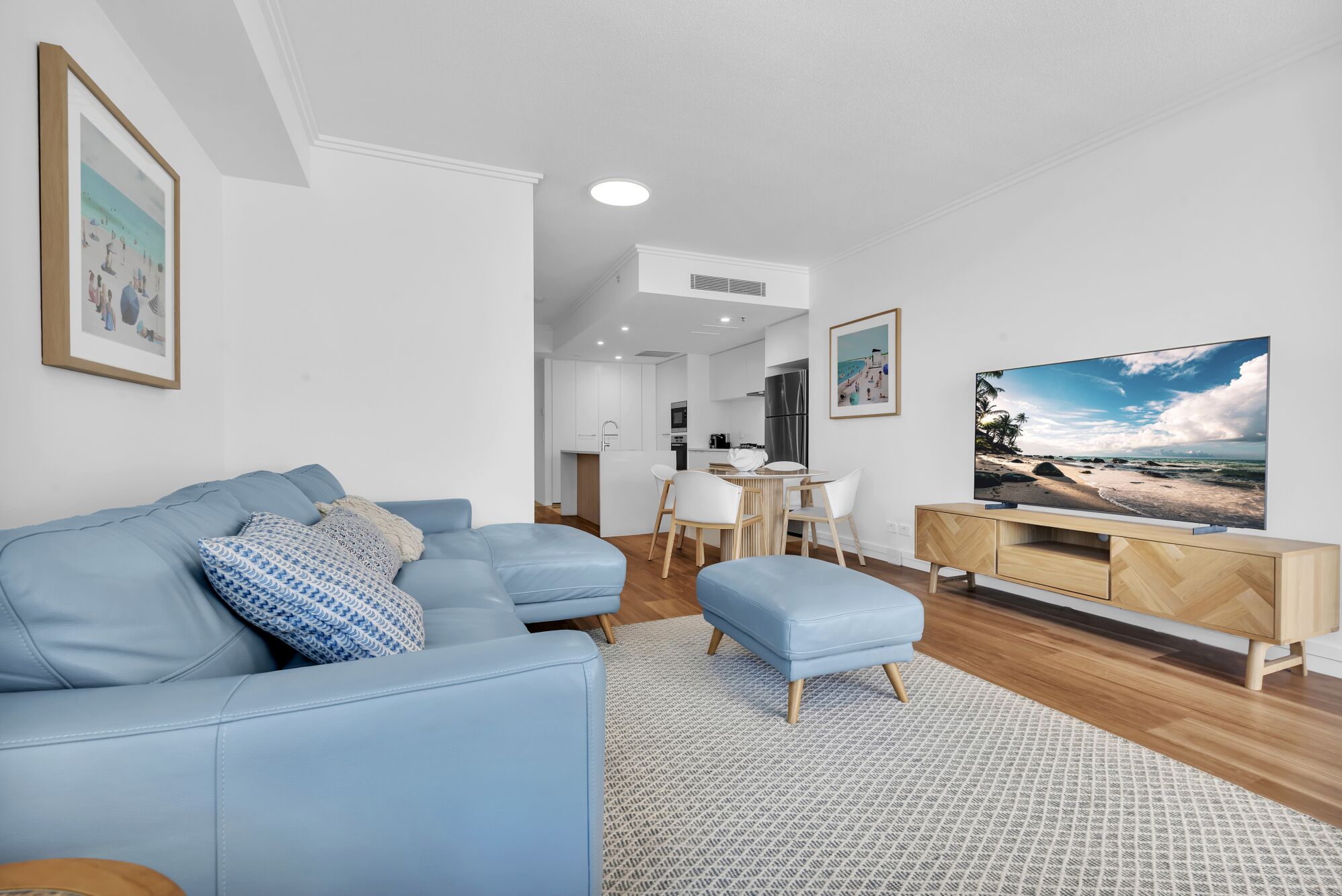 A modern living room with a blue sofa, a dining table with chairs, a TV on a wooden stand, and a kitchen in the background.
