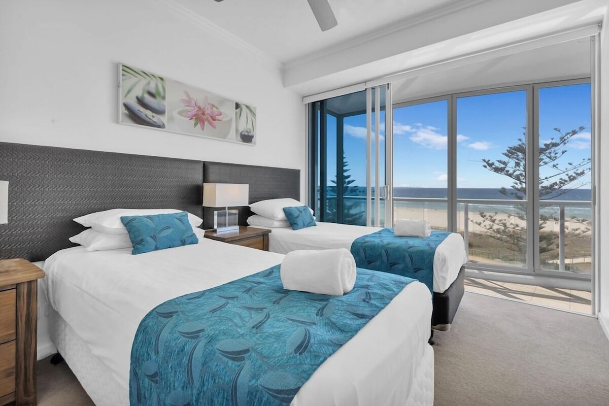 A hotel room with two beds, a large window with a view of the beach, and a ceiling fan.