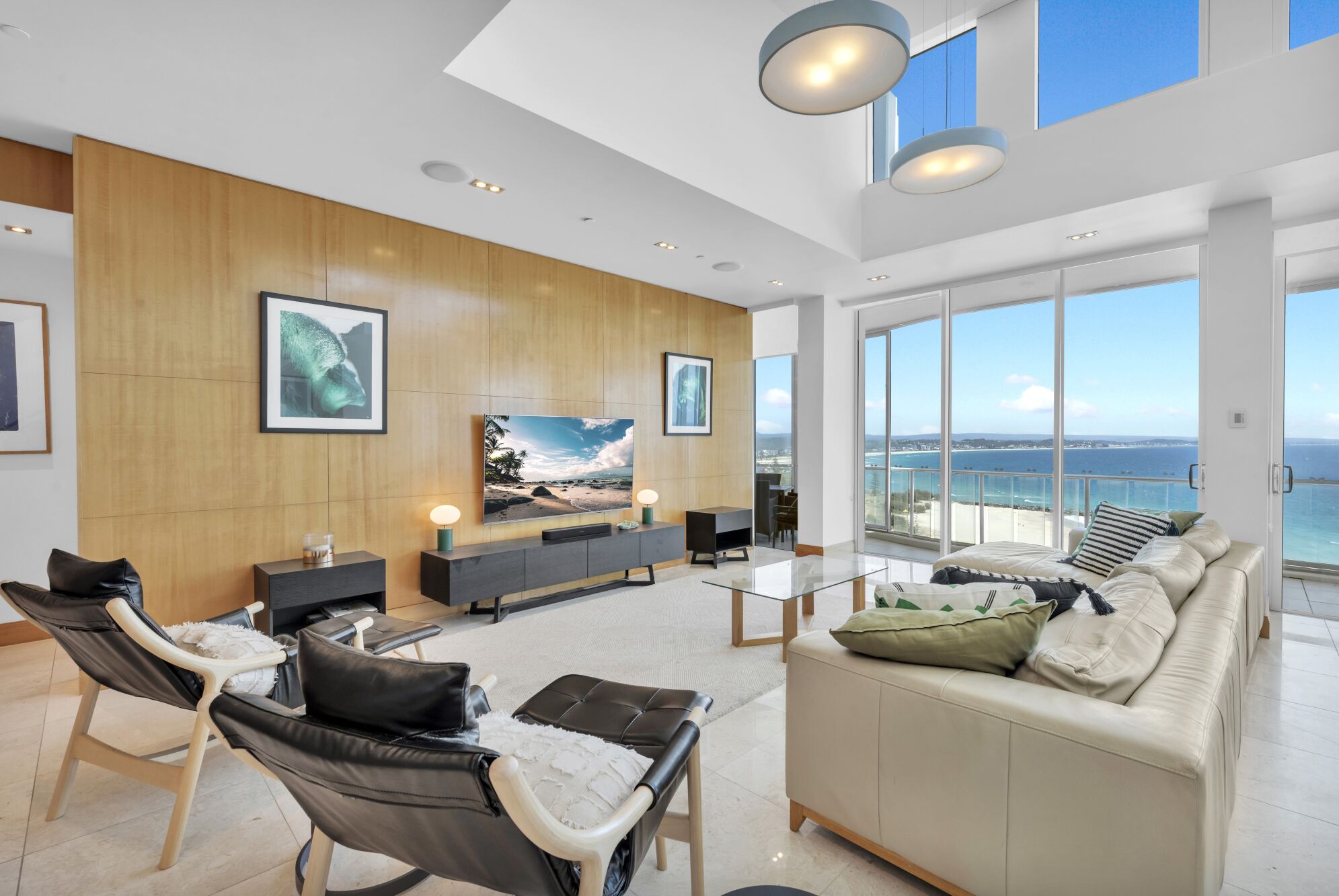 A spacious living room with a large TV, comfortable seating, wooden panel walls, and a balcony with a sea view.