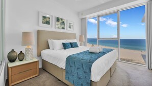 A bedroom with a bed, bedside lamps, a nightstand, and a large window with a beach view. - Reflection Tower Two (Coolangatta)