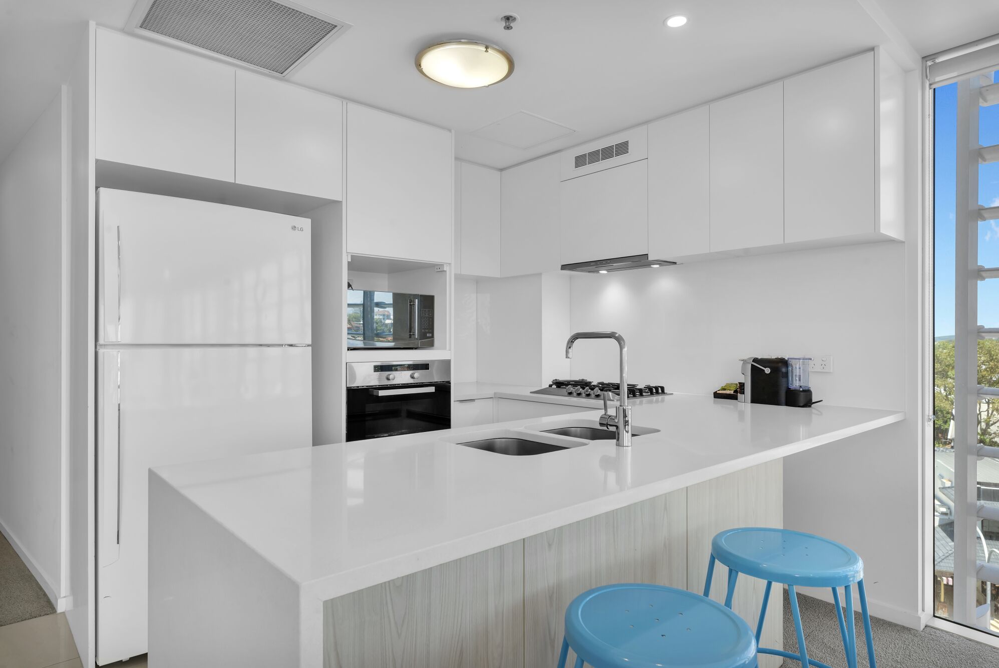 A modern kitchen with white cabinetry, a built-in microwave, a stainless steel sink, and a gas stove. There are two blue stools and a window providing a view of the outdoors.
