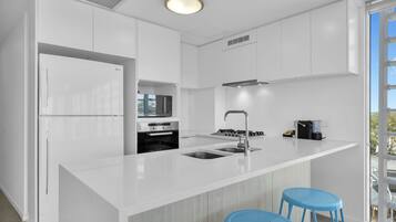 A modern kitchen with white cabinetry, a built-in microwave, a stainless steel sink, and a gas stove. There are two blue stools and a window providing a view of the outdoors.