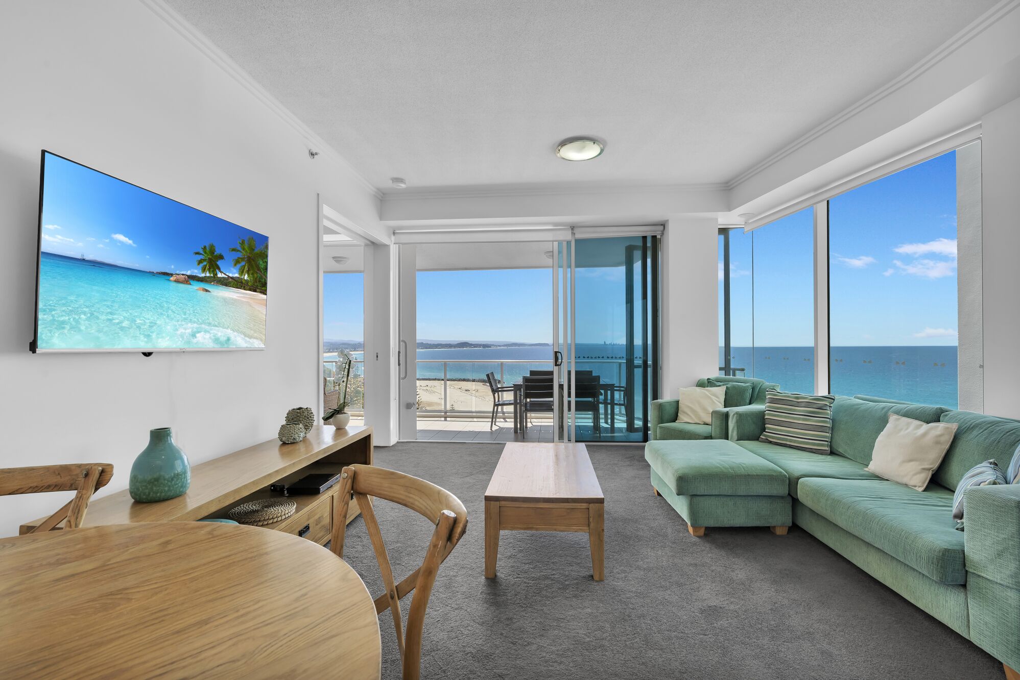 A spacious living area with a large flat-screen TV, a green sofa, a wooden coffee table, and a dining table with chairs.